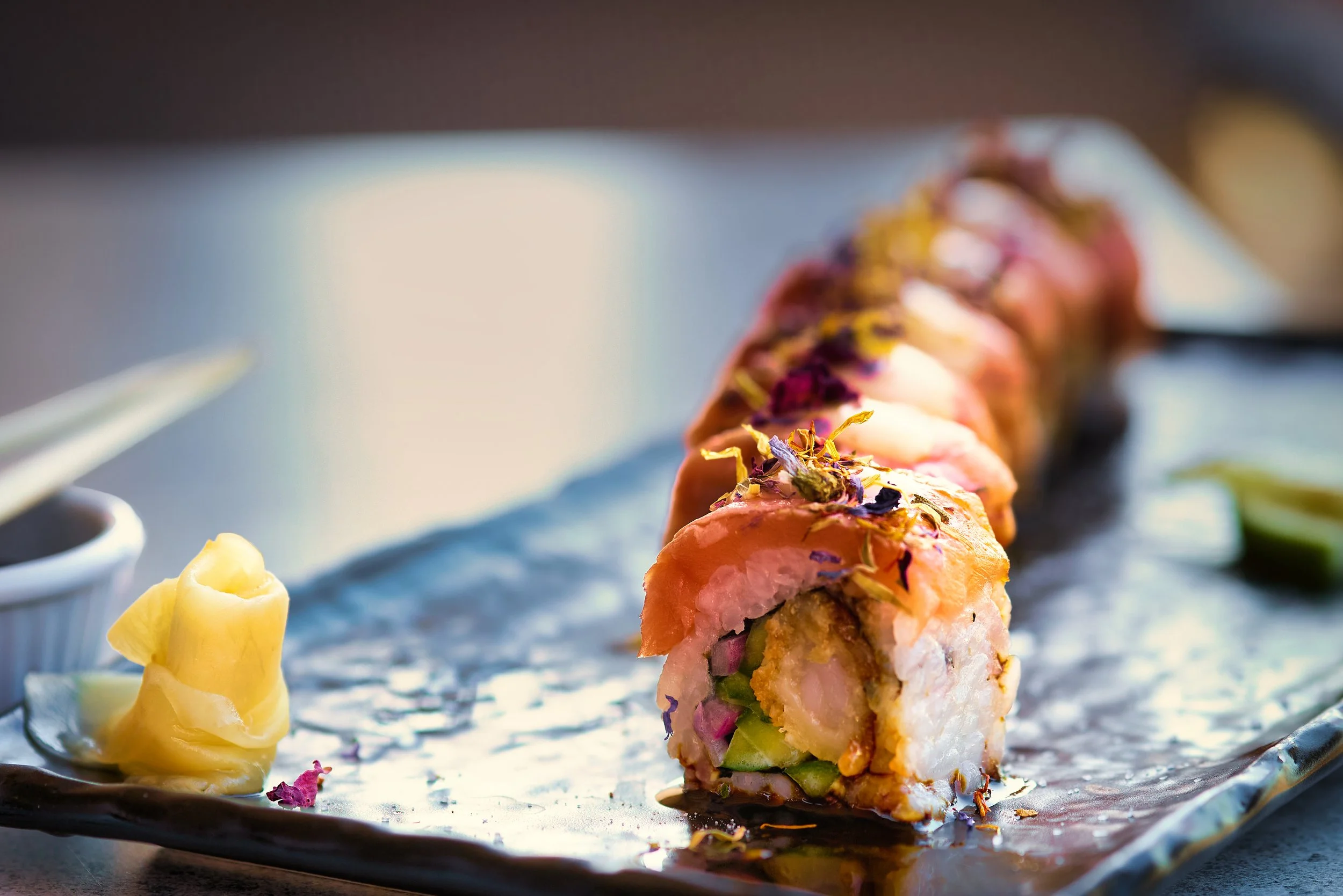 Close-up of a sushi roll garnished with edible flowers on a black plate, beside a dollop of yellow wasabi.