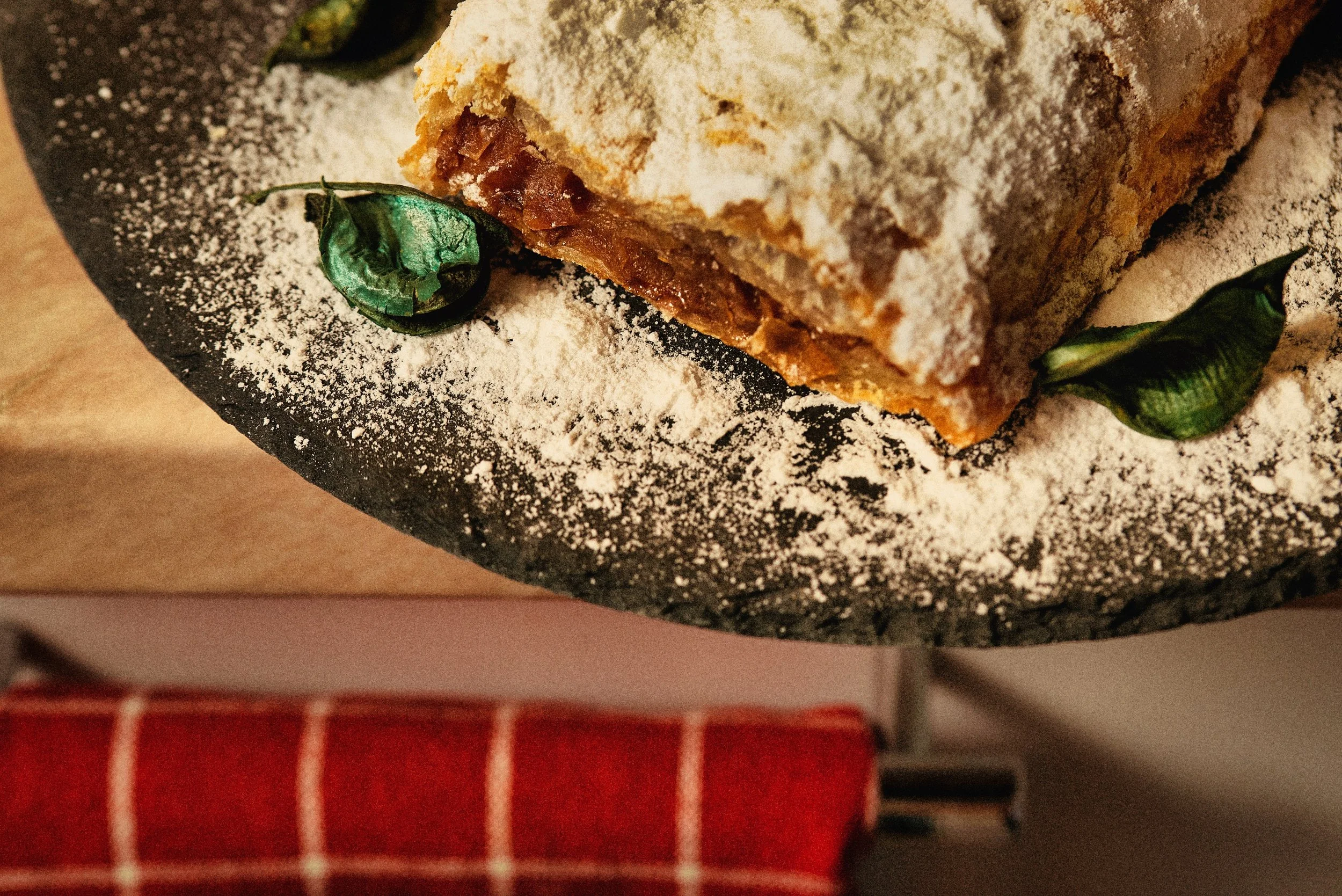 A slice of fruit-filled pie topped with powdered sugar, placed on a black serving dish with powdered sugar dusted around it and green decorative leaves.