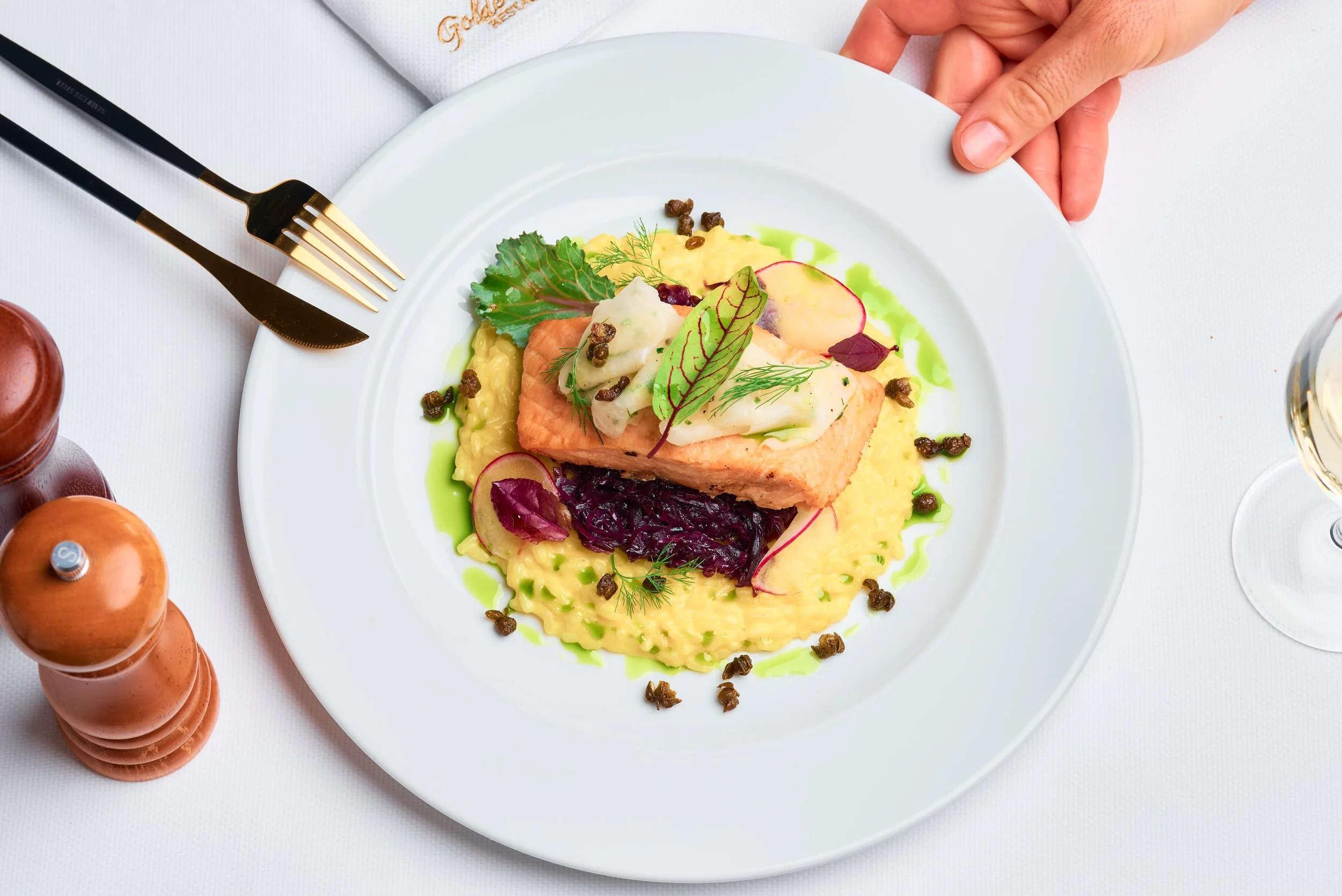 A plated dish consisting of a piece of cooked salmon topped with sauce, garnished with herbs and microgreens, served with mashed potatoes and vegetables, on a white plate with black and gold fork, on a white tablecloth.