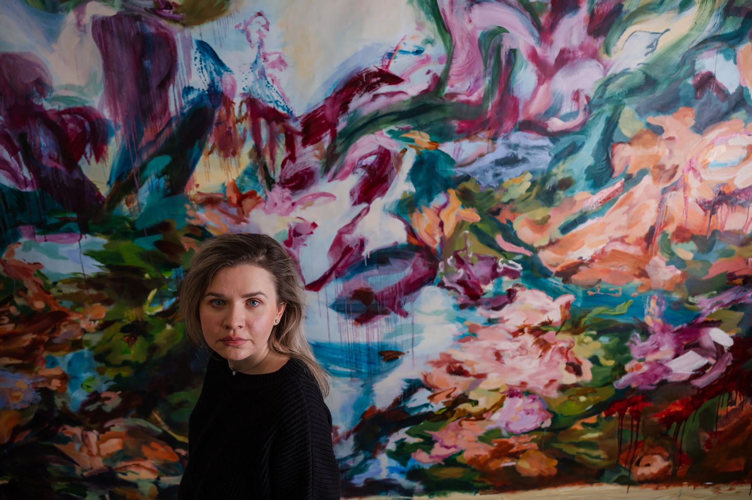 A young woman with light brown hair and a black sweater standing in front of a colorful abstract painting with swirling brushstrokes of pink, green, blue, orange, and white.