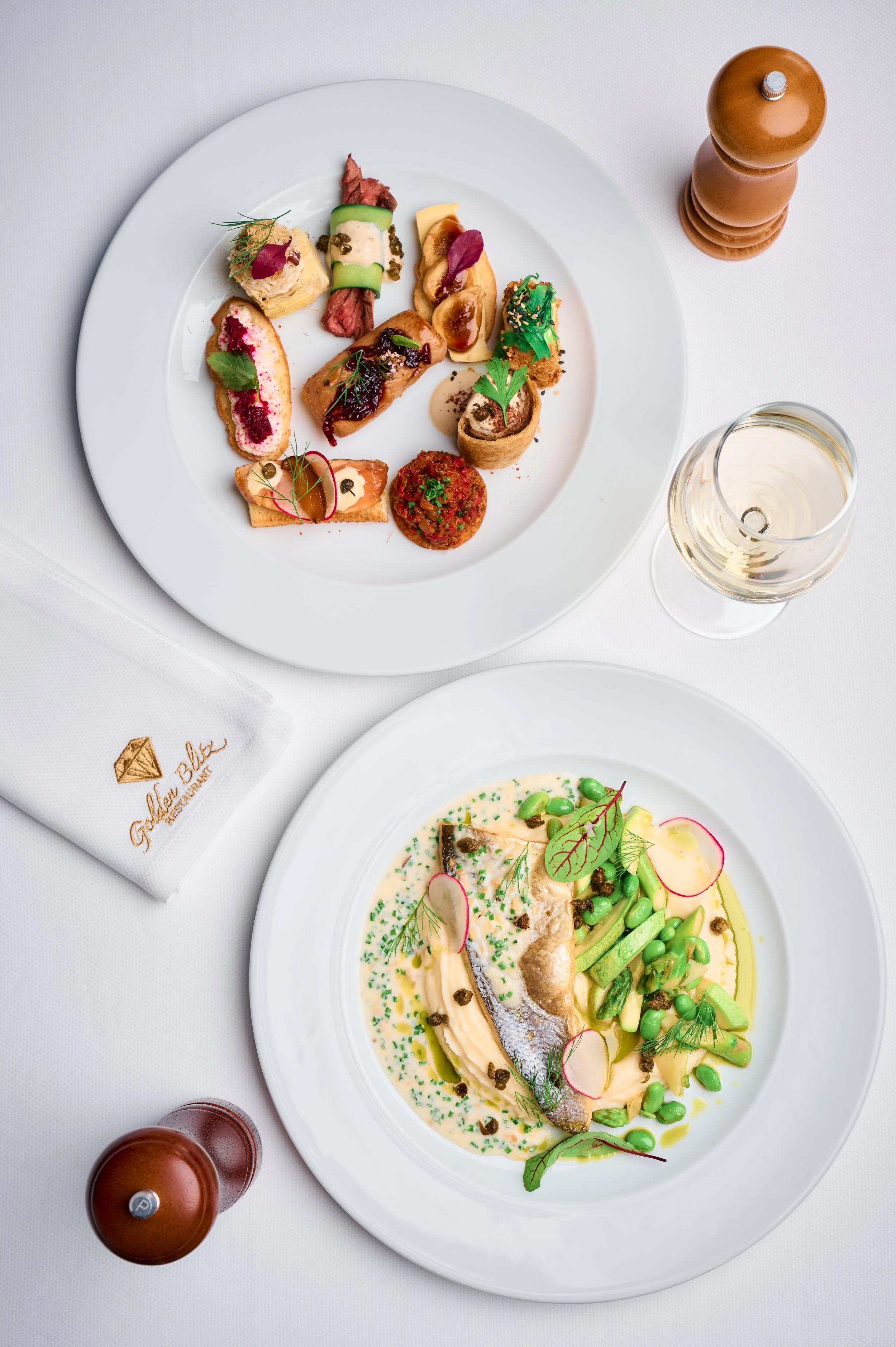 A white plate with small, artistically arranged appetizers and microgreens, next to a glass of white wine and salt and pepper shakers on a white tablecloth.