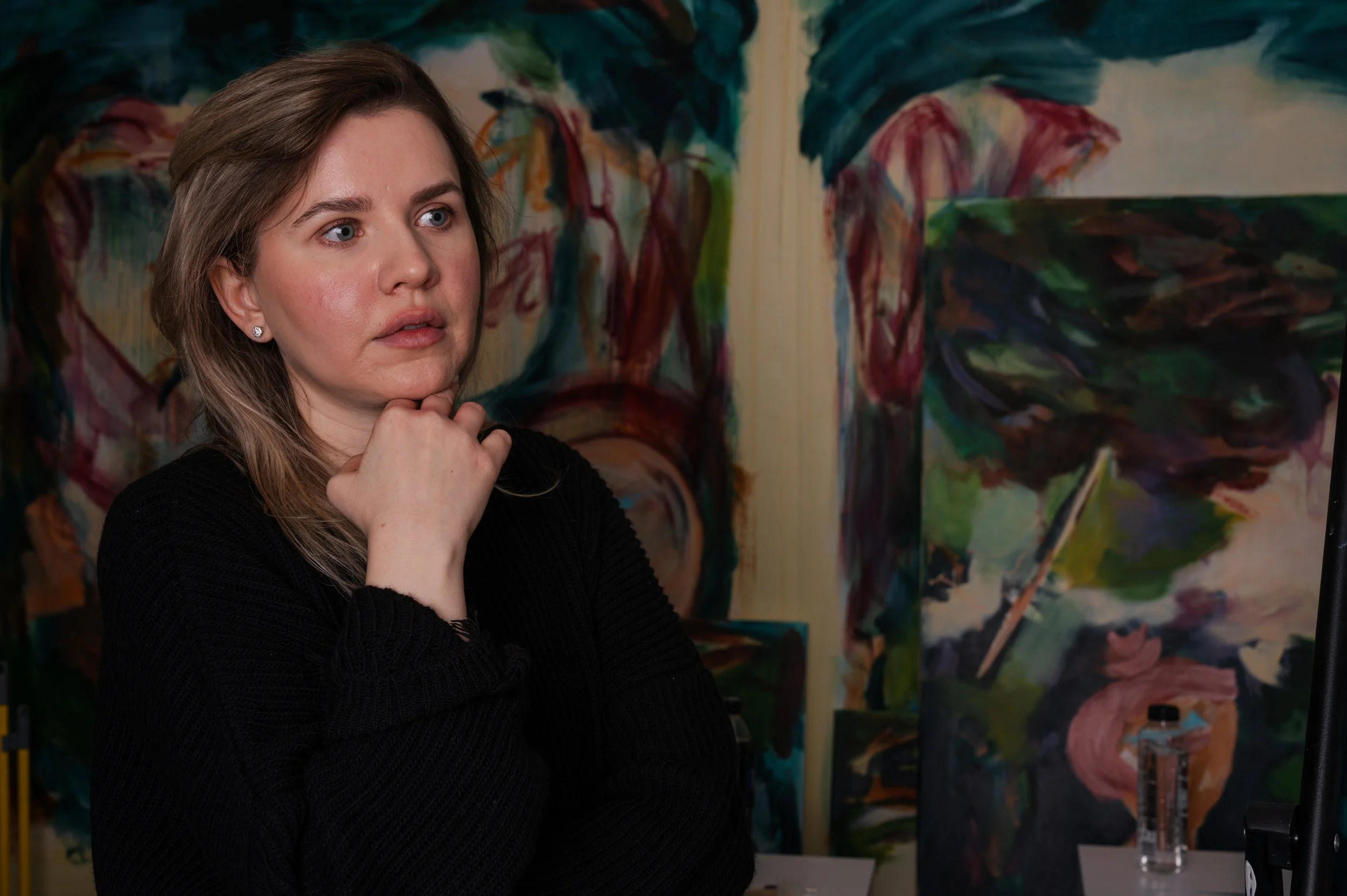 A woman with brown hair and blue eyes looking at a colorful abstract painting, with her hand resting on her chin.