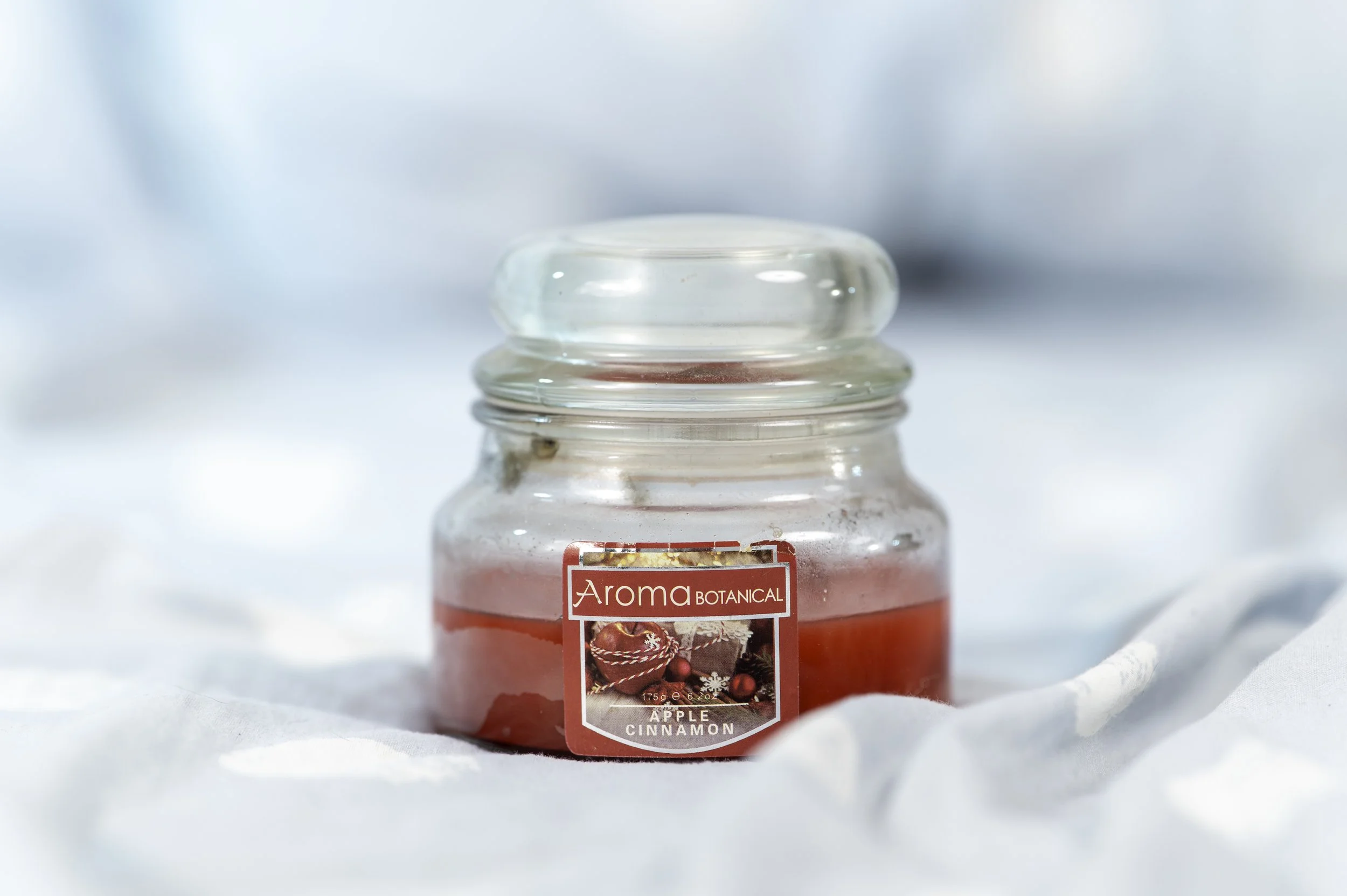 A clear glass jar with a red and white label reading 'Aroma Botanical' and 'Apple Cinnamon', containing a candle or scent product, placed on a white surface with a soft, light blue background.