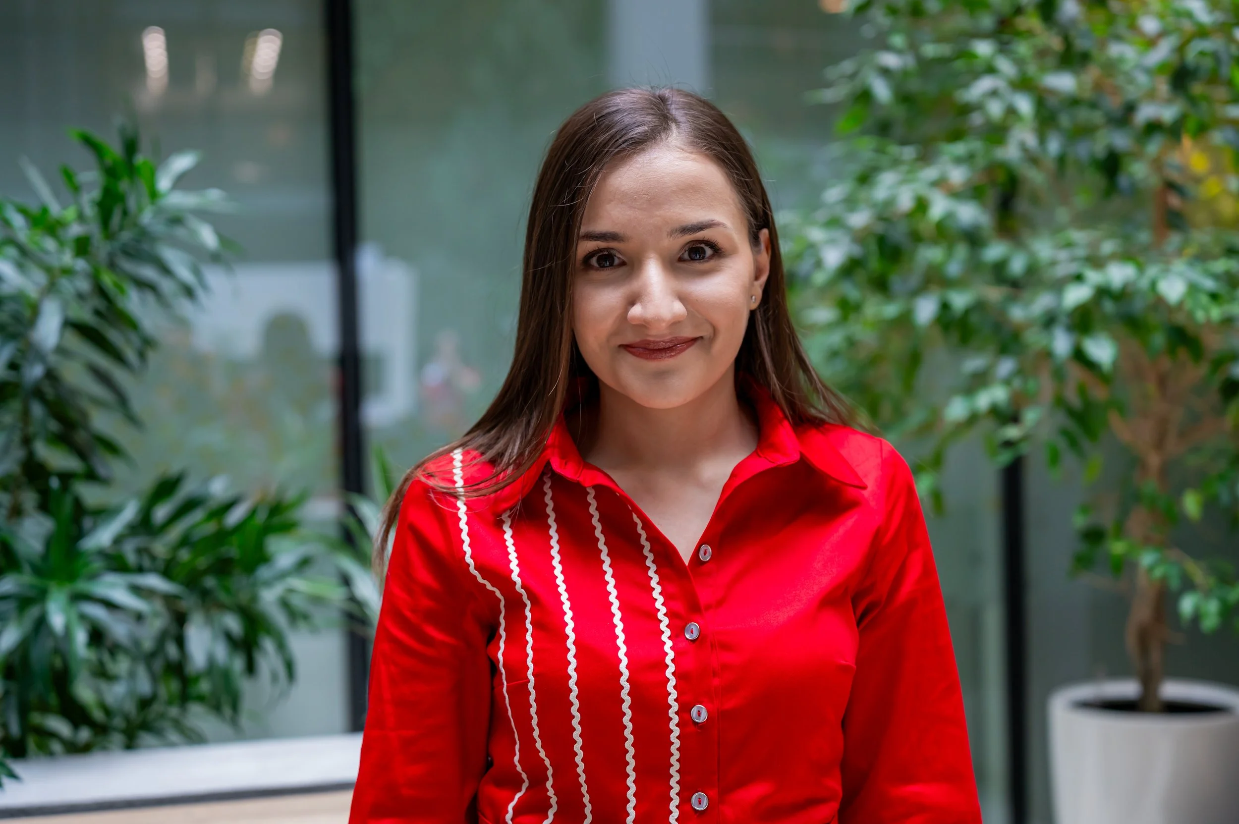 A woman with long brown hair wearing a red shirt with white stripes, standing indoors near large plants and glass windows.