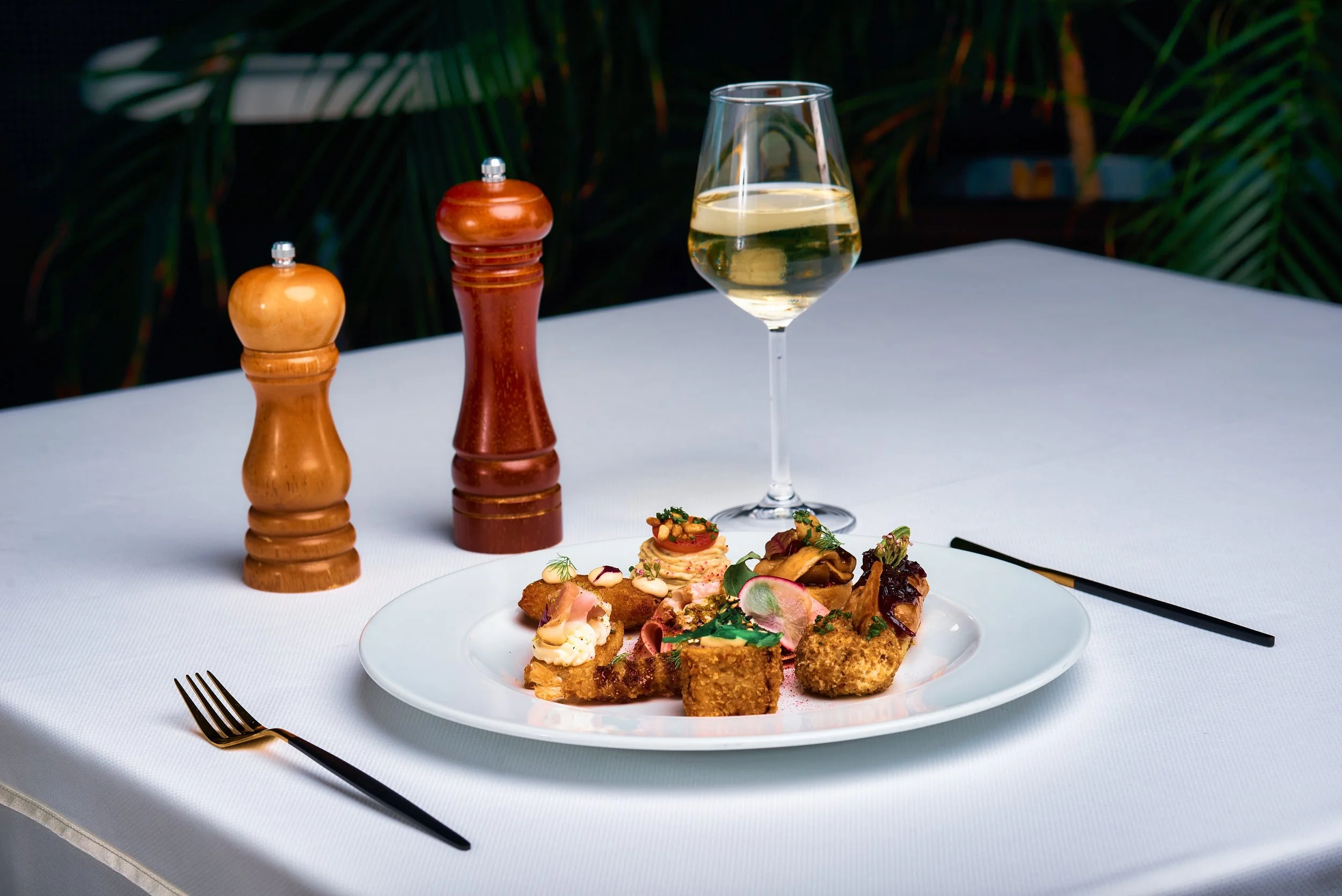 A white table with a white plate filled with various fried and garnished foods, a glass of white wine, and salt and pepper shakers in the background, with greenery behind.