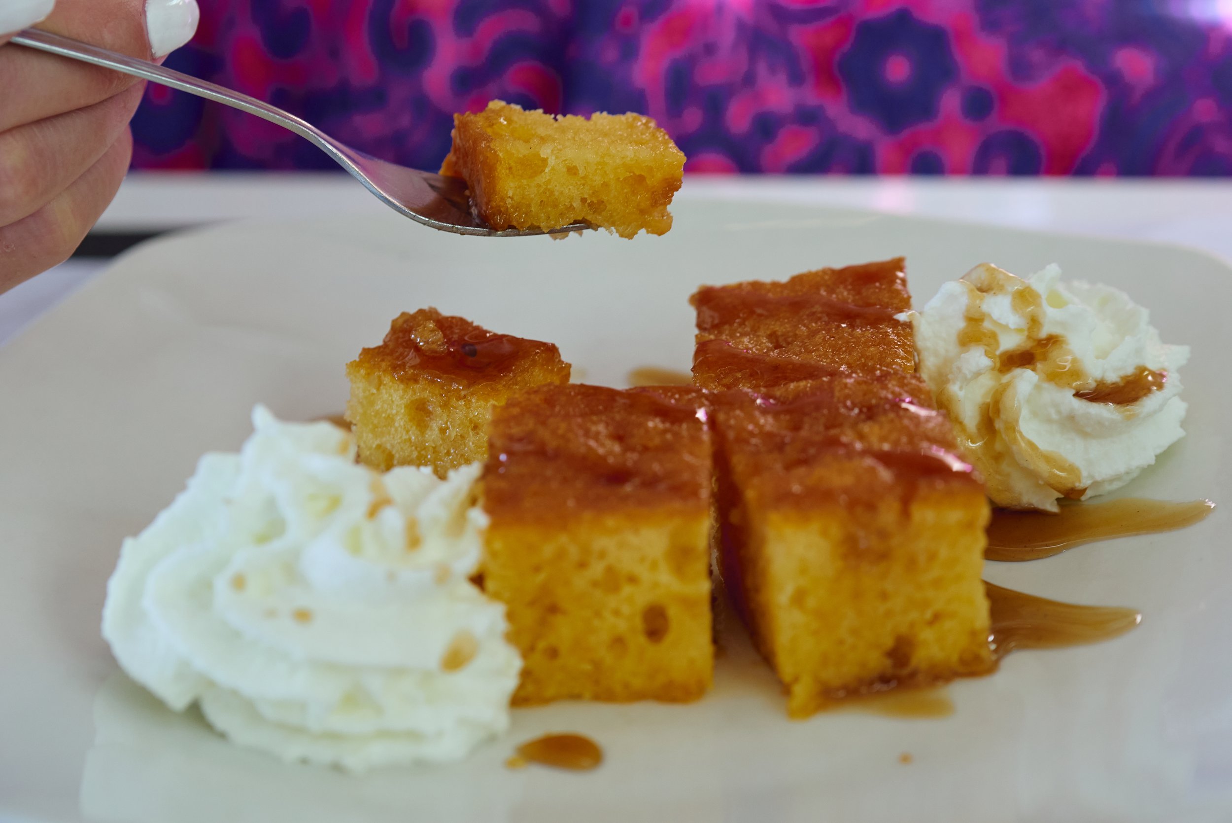 Close-up of a fork holding a piece of golden yellow cornbread above a plate with more pieces of cornbread, mashed potatoes with gravy, and whipped cream.