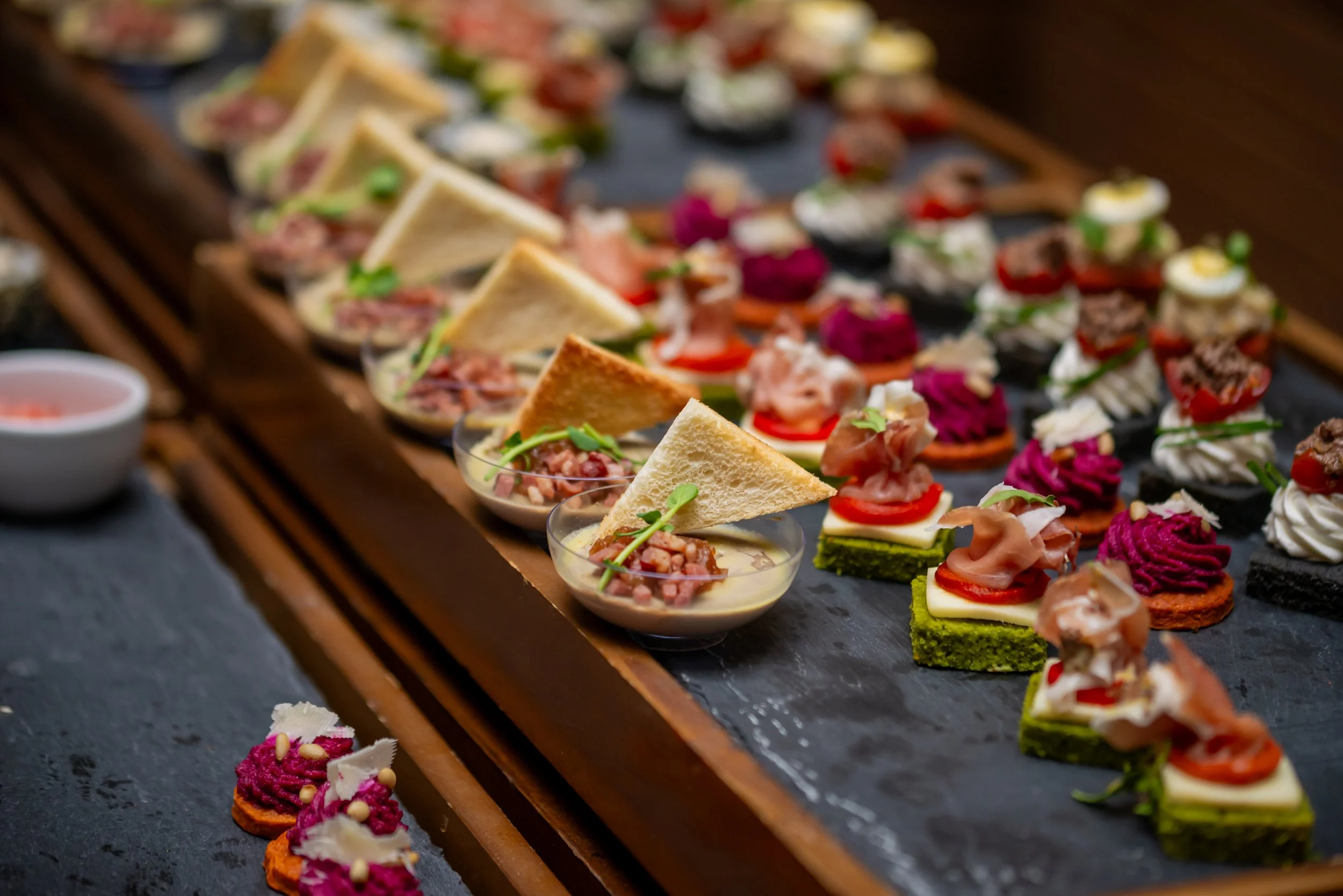 Assorted gourmet appetizers on a platter, including small sandwiches, cheese on crackers topped with meats and vegetables, and other bite-sized items.