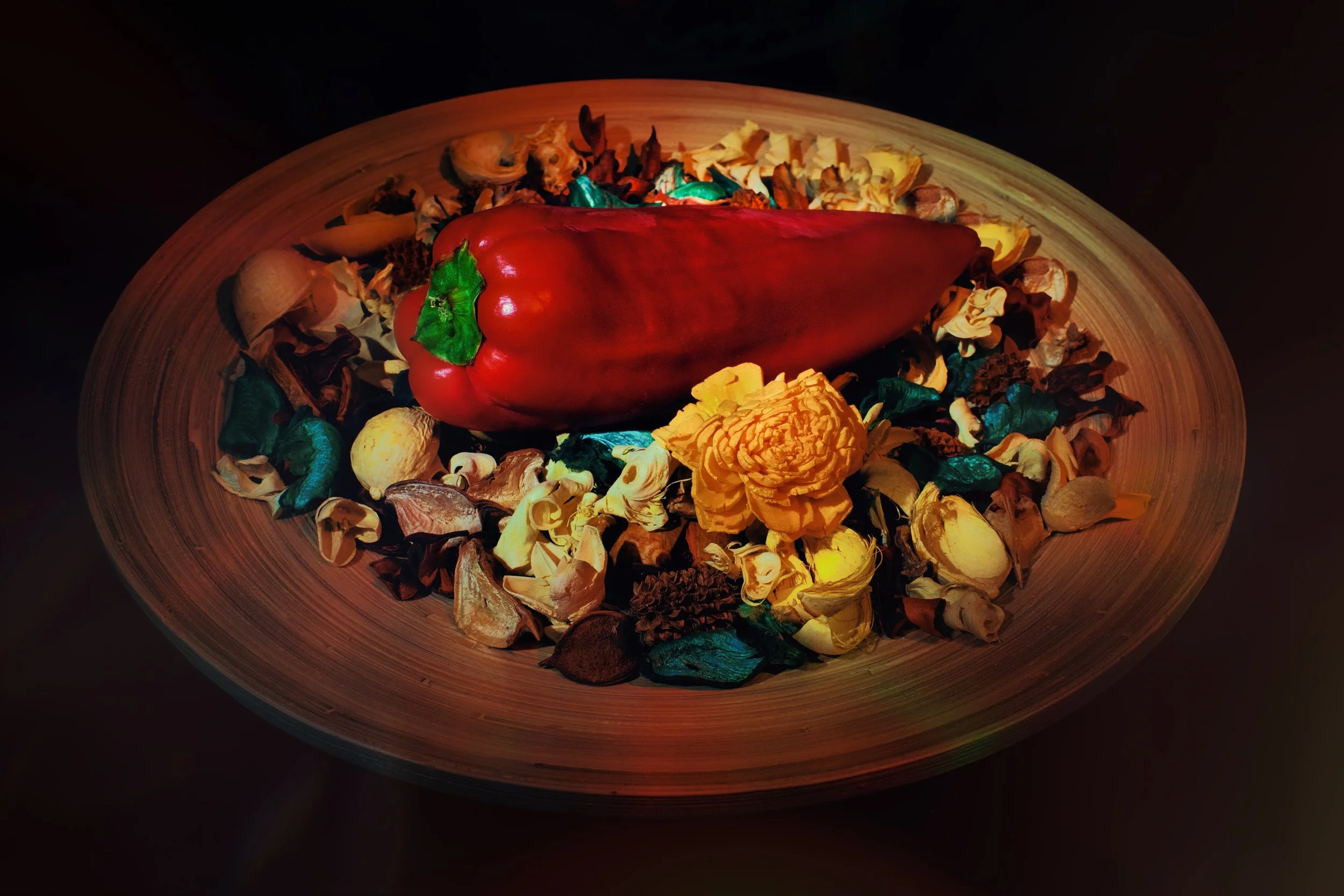 A large red bell pepper resting on a round wooden tray surrounded by dried flower petals and potpourri.
