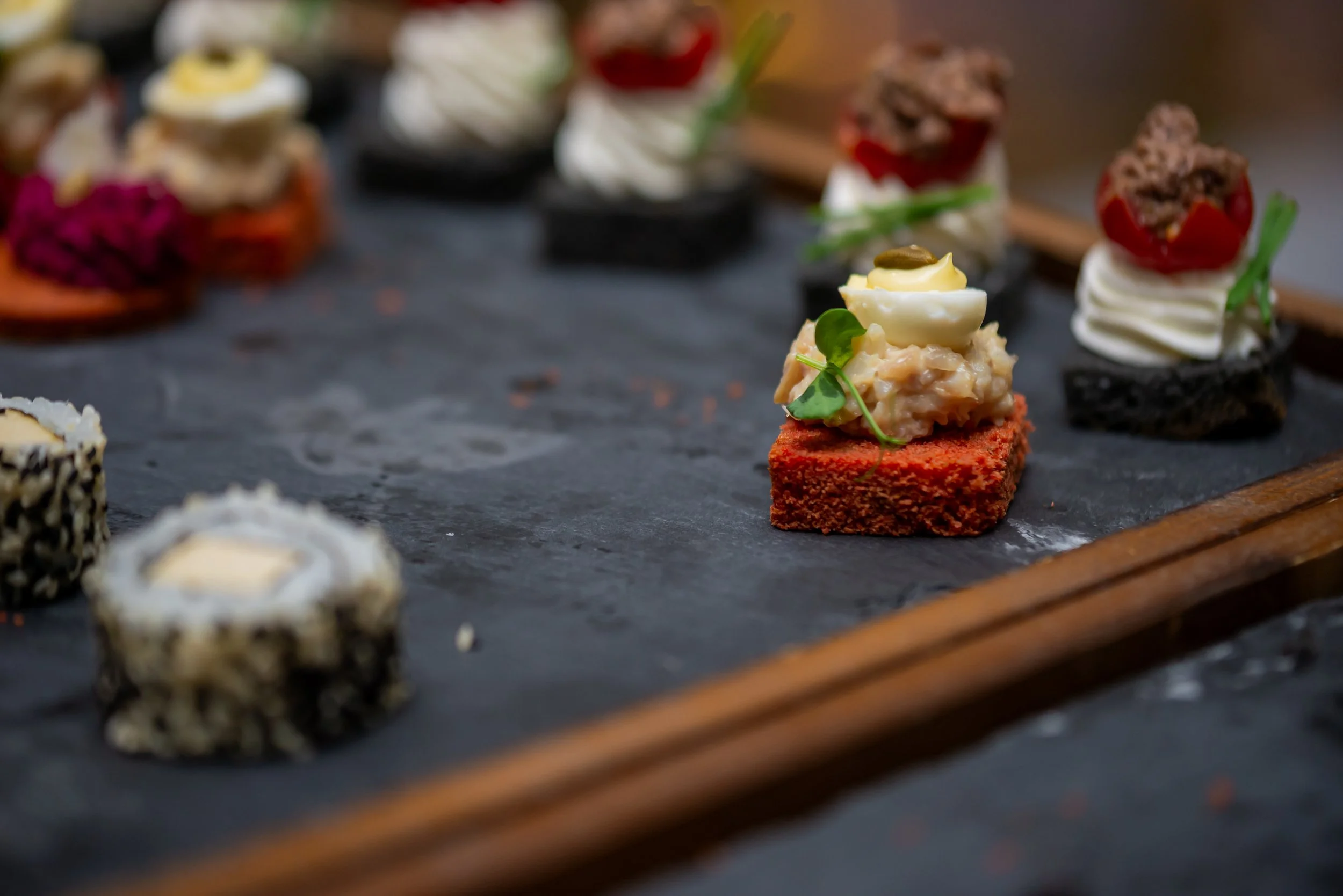 Assorted gourmet appetizers on a black slate serving board, featuring bite-sized pieces with various toppings including egg, vegetables, and sauces.