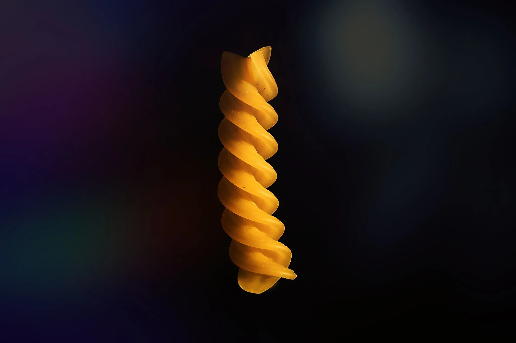 Close-up of a yellow, spiral-shaped pasta against a dark background with colorful light effects.