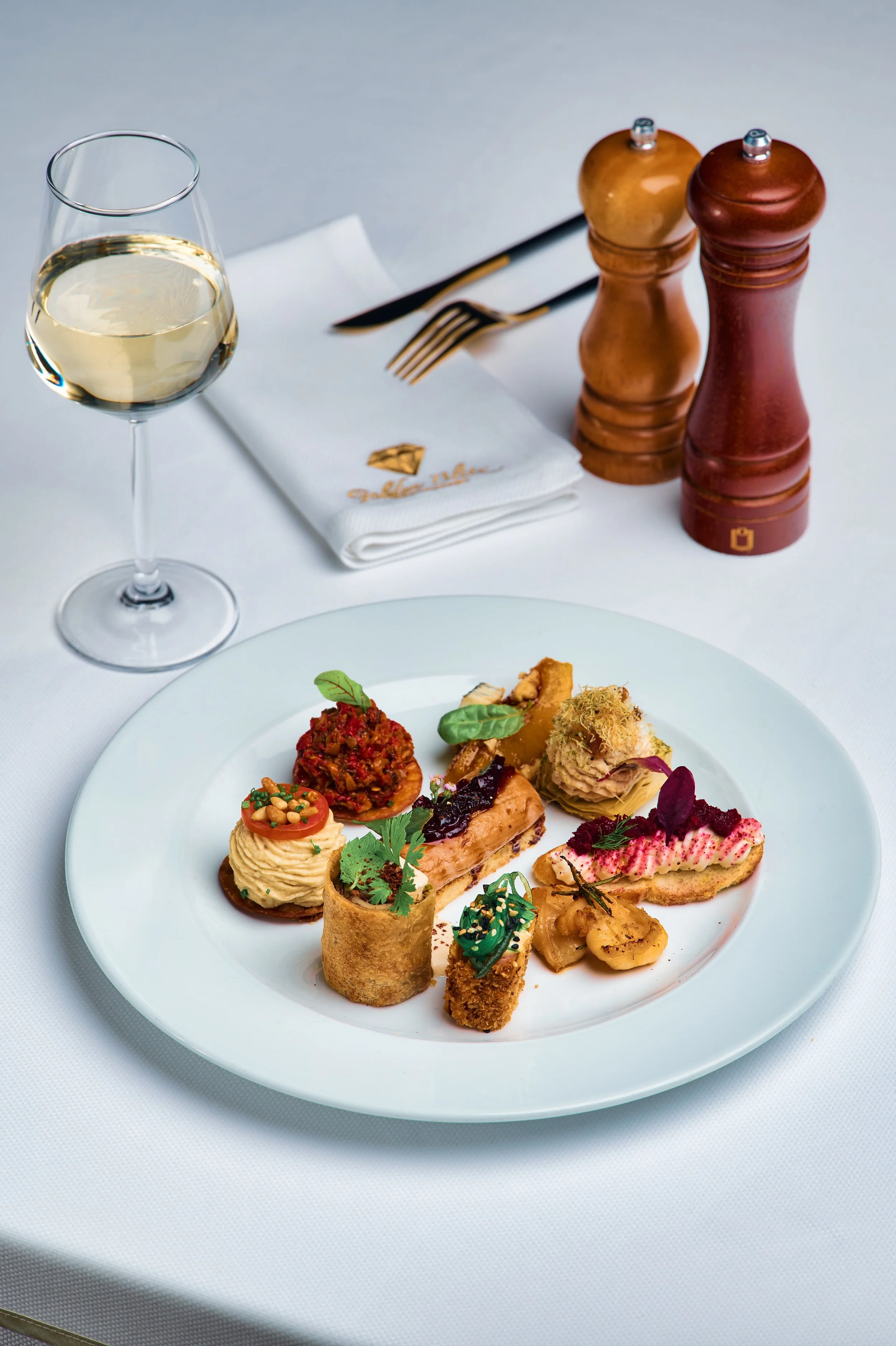 A white plate with various gourmet appetizers and small bites, including small rolls, mousse, various toppings, garnishes, and sauces, positioned on a white tablecloth. In the background, there is a glass of white wine, a white cloth napkin, a fork, 