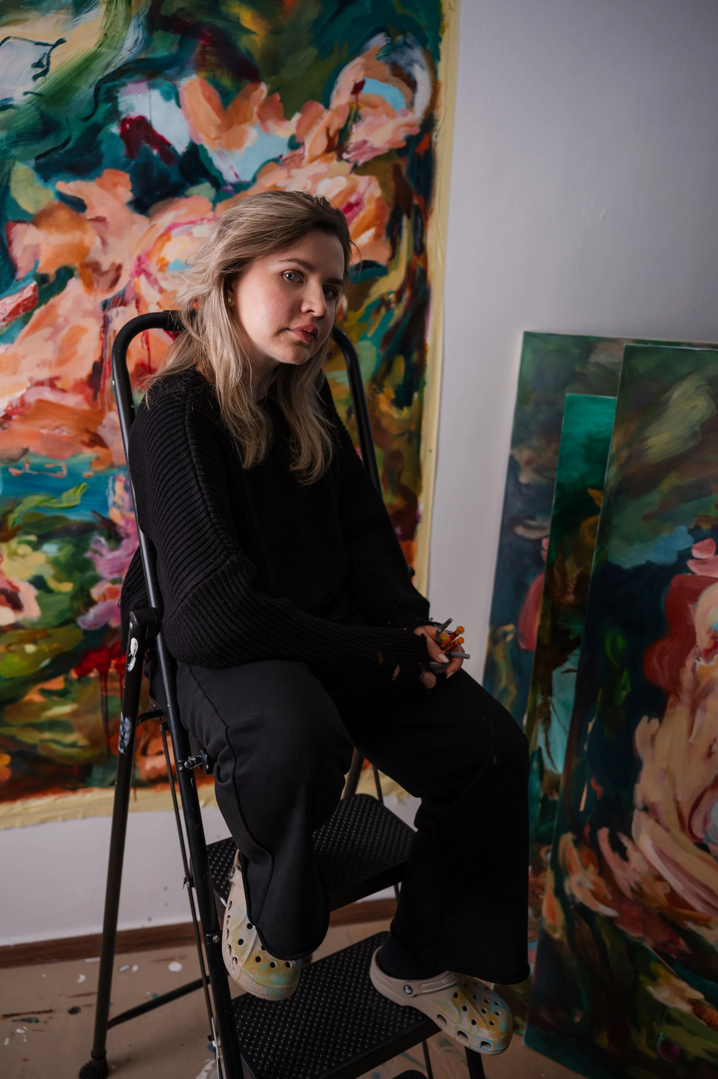 A woman with shoulder-length blonde hair and light skin sitting on a black chair in an art studio, surrounded by colorful abstract paintings. She is wearing a black sweater, black pants, and yellow shoes with a floral pattern, holding a paintbrush an