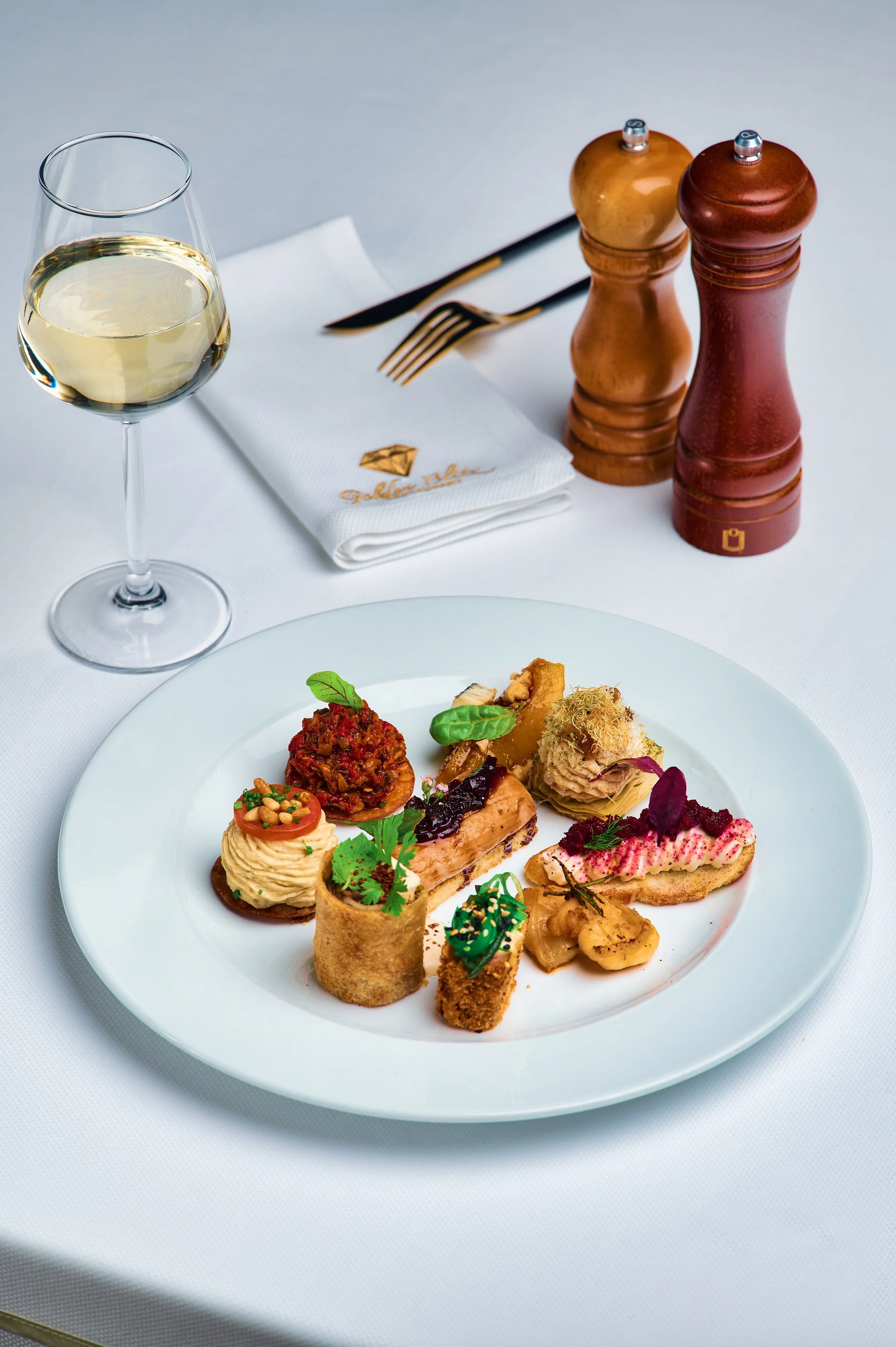 A white plate with a variety of gourmet appetizers and finger foods including small bites of meat, vegetables, and decorative garnishes, on a white tablecloth. In the background, there is a glass of white wine, salt and pepper shakers, a fork, and a 