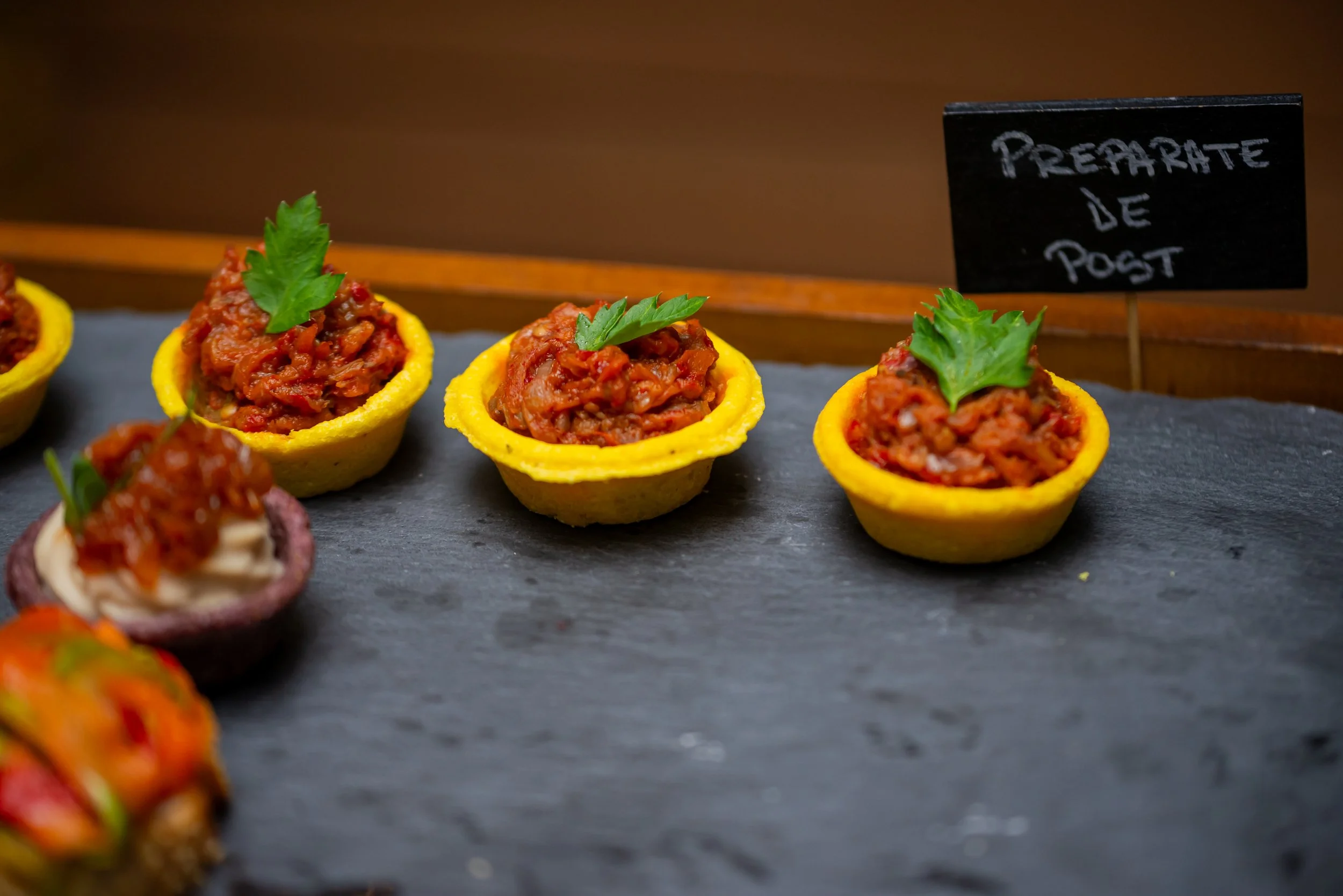 Small yellow taco cups filled with seasoned minced meat topped with green parsley leaves, displayed on a dark slate serving platter with a handwritten sign in the background that reads 'Prepararte de Post'.