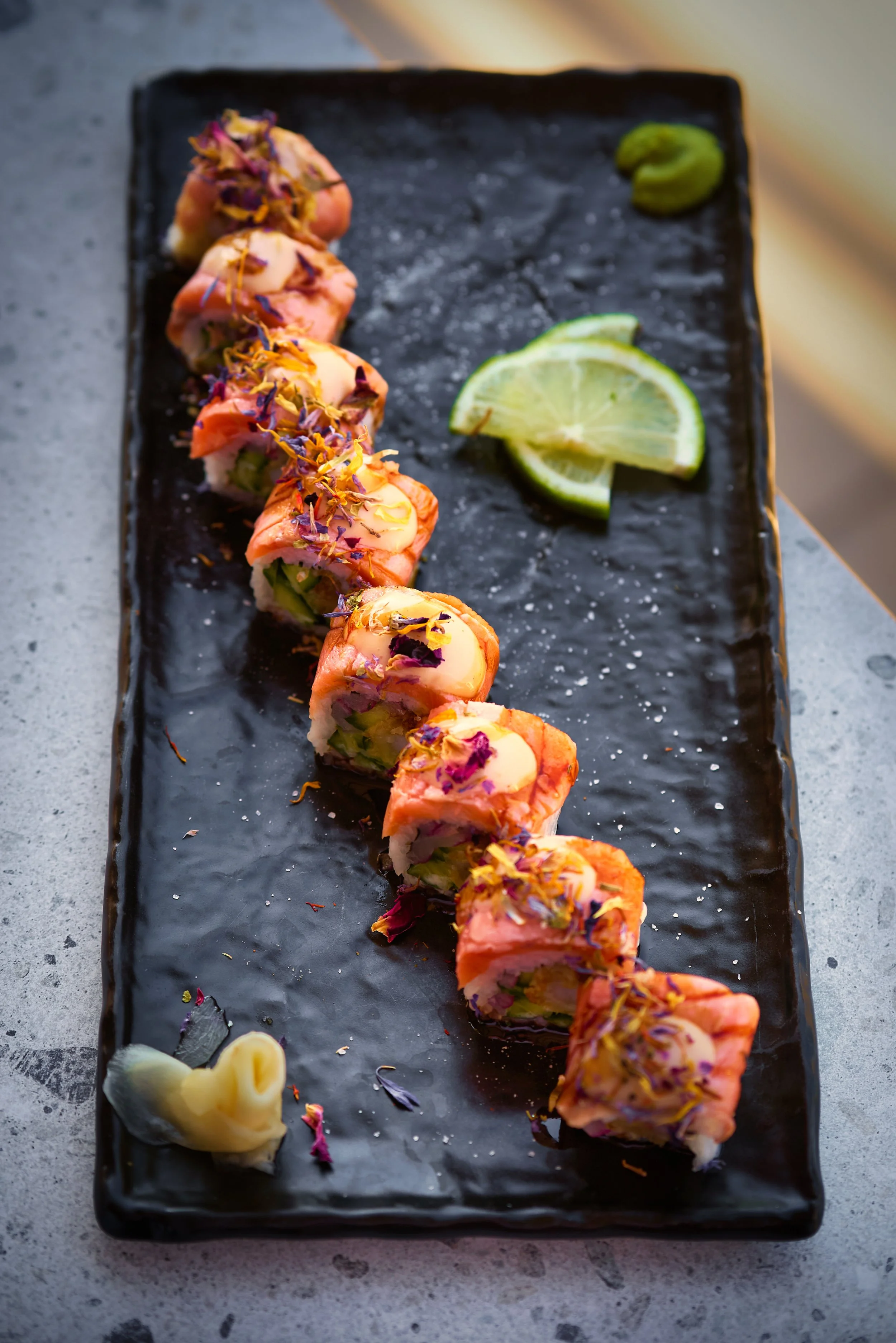 Sushi rolls with colorful toppings on a long black rectangular plate, garnished with lime wedges, wasabi, and pickled ginger.