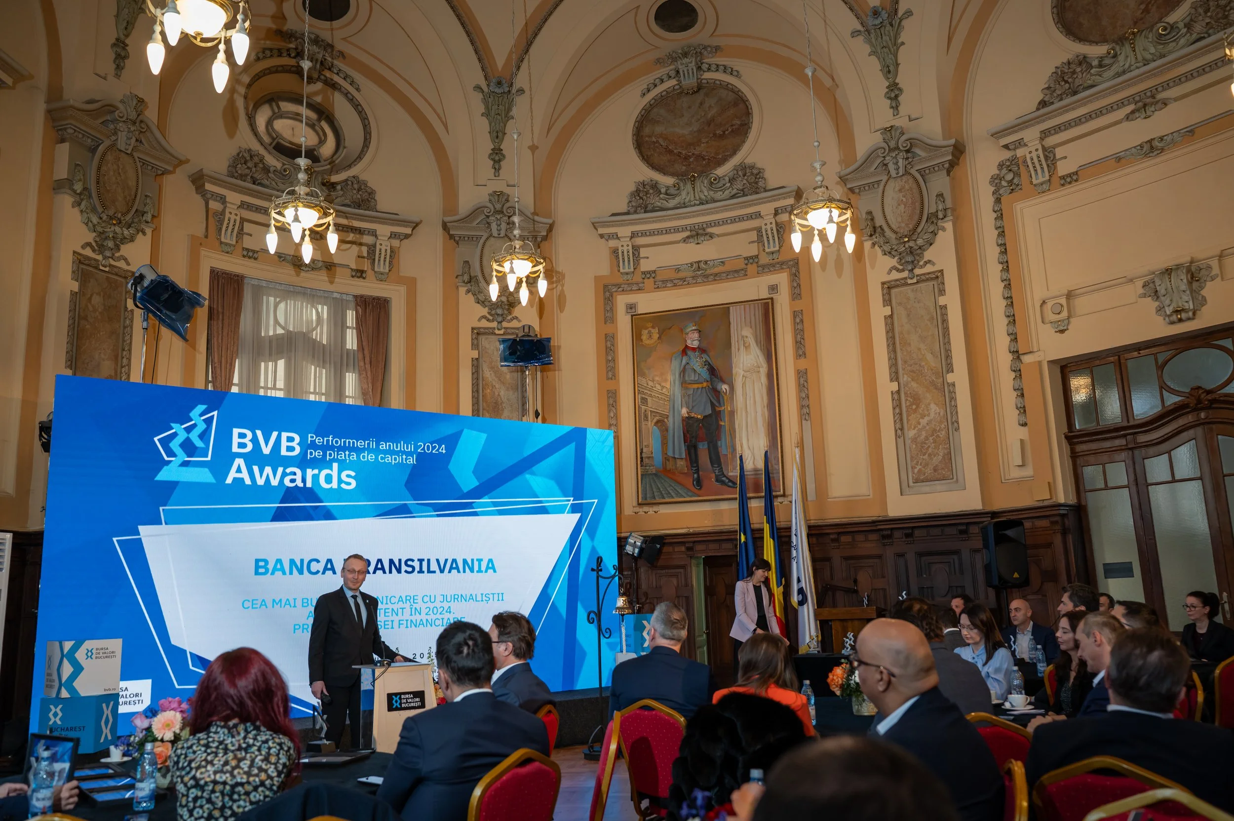 A conference or awards ceremony taking place in a grand, ornately decorated room with chandeliers, murals, and high ceilings. There is a large screen displaying the event title and a speaker at a podium.