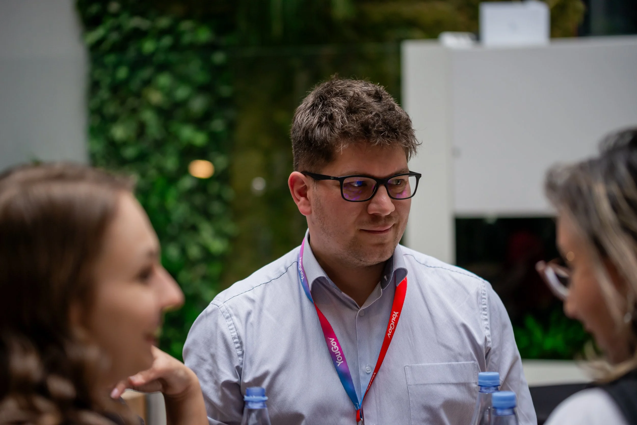 A man with glasses and a lanyard engaged in conversation at an outdoor event, with blurred people and greenery in the background.