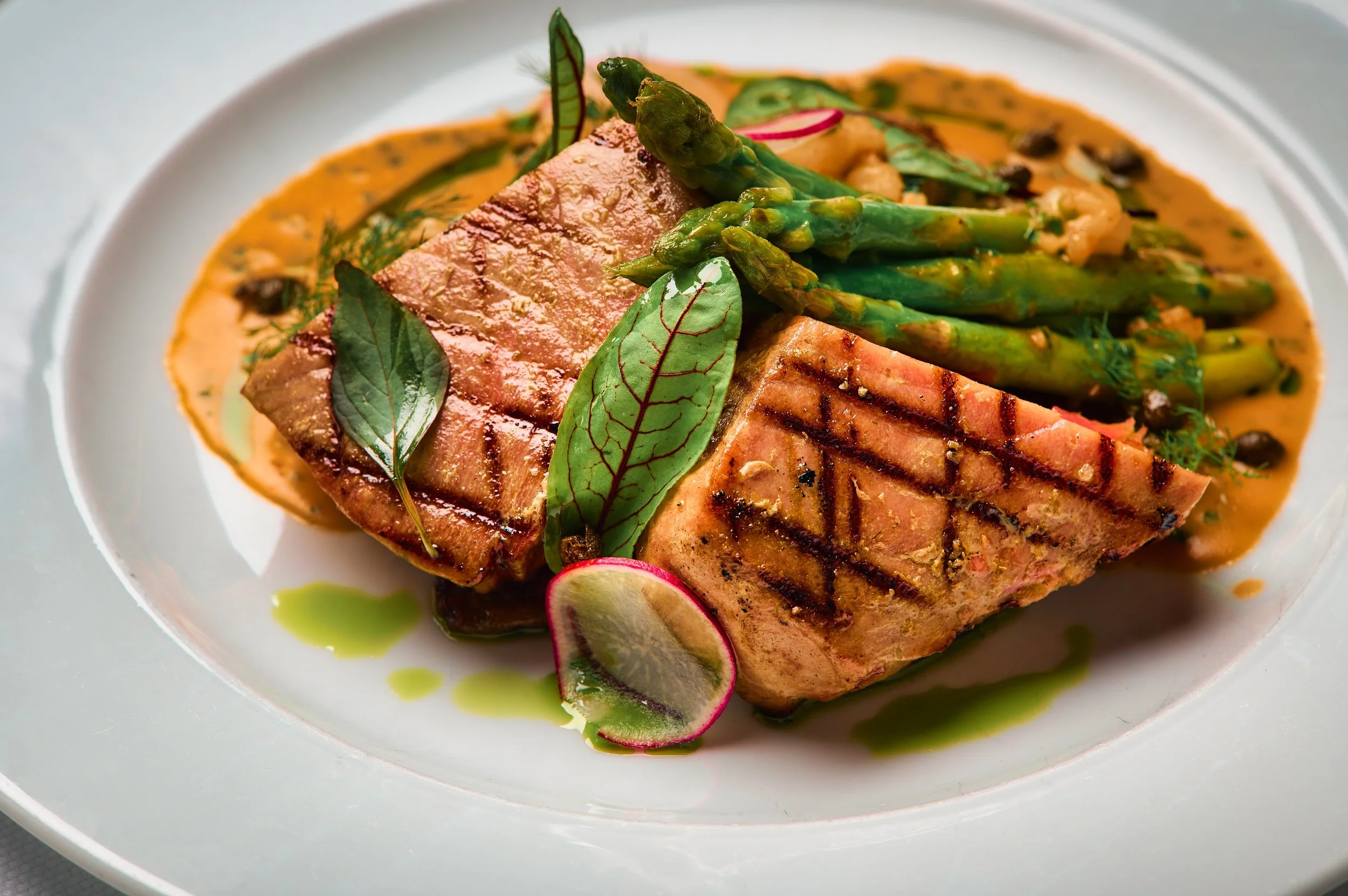 A plate of grilled salmon topped with asparagus spears, radish slices, and fresh herbs, with a drizzle of green sauce around the plate.