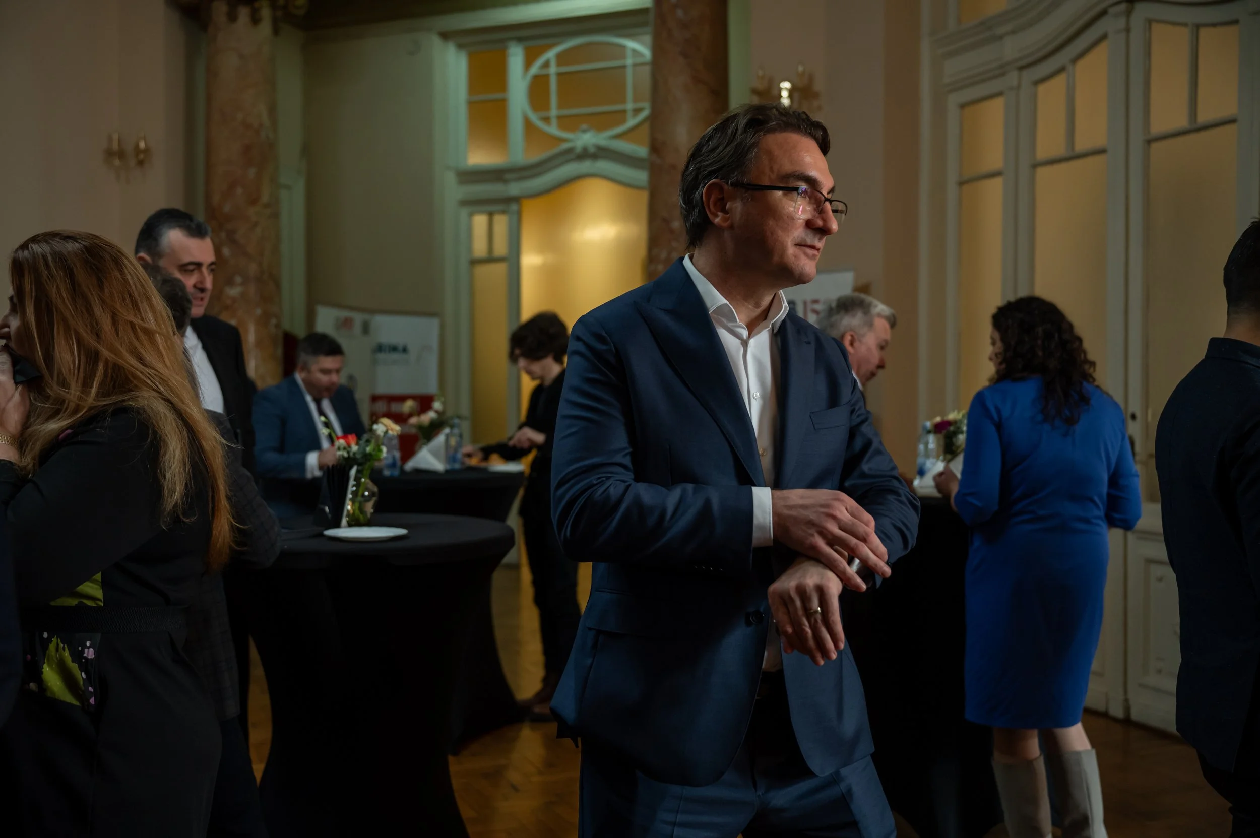 A man in a blue suit and glasses standing at a formal gathering or event, surrounded by other people in professional attire, in a decorated indoor venue with ornate walls and floral arrangements.