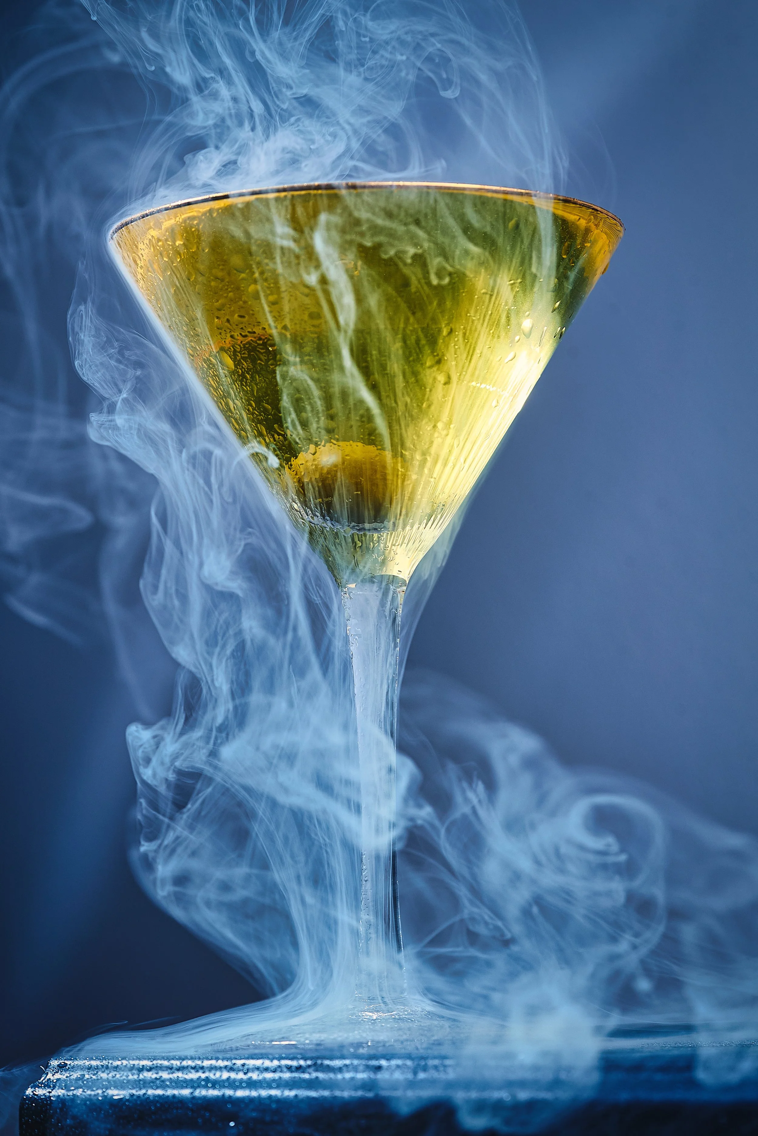 A close-up of a martini glass filled with a yellow-colored drink, with smoke or vapor swirling around and above the glass against a dark background.