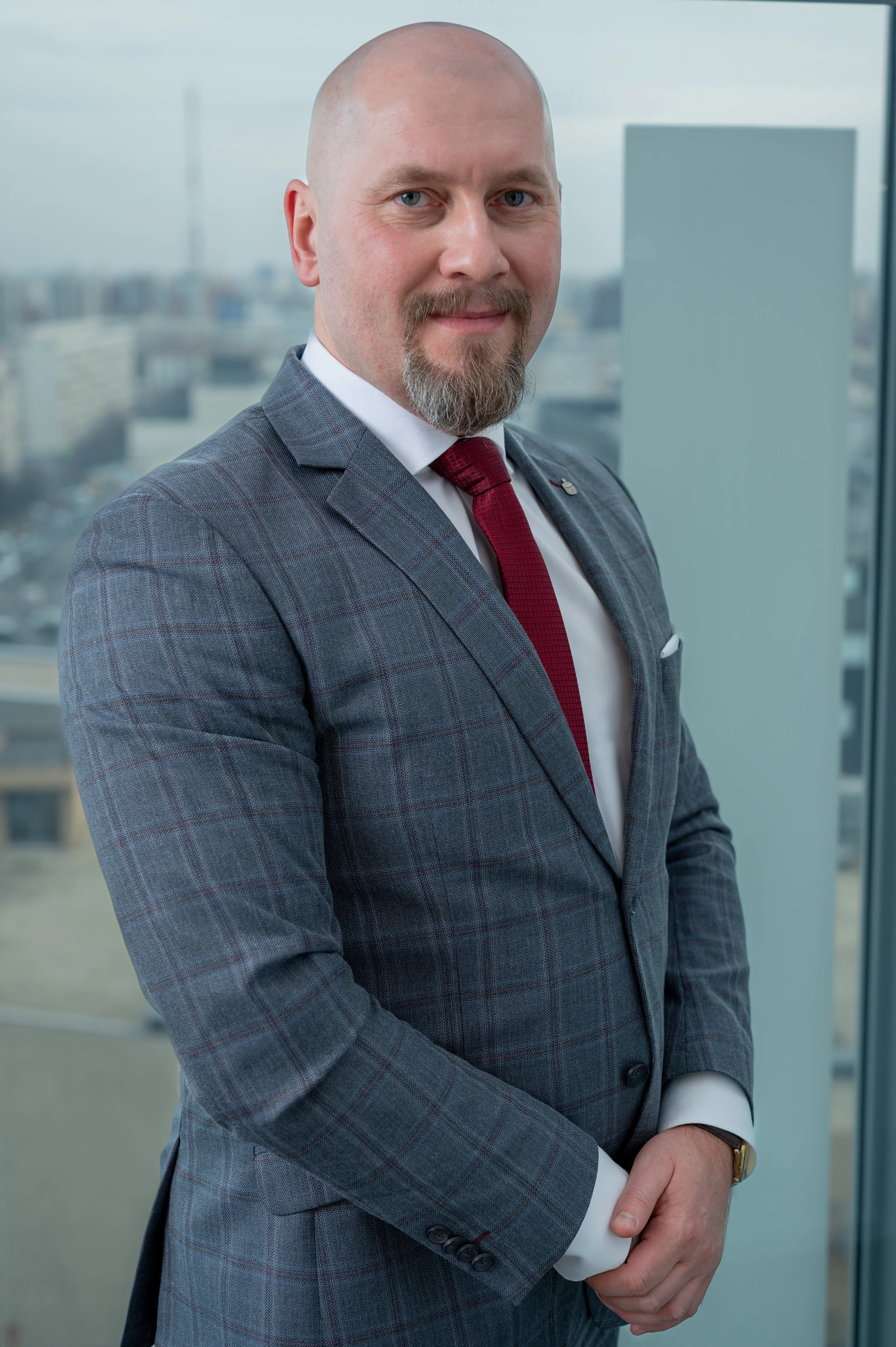 A man in a checked suit with a red tie standing indoors, with a cityscape view through a large window behind him.