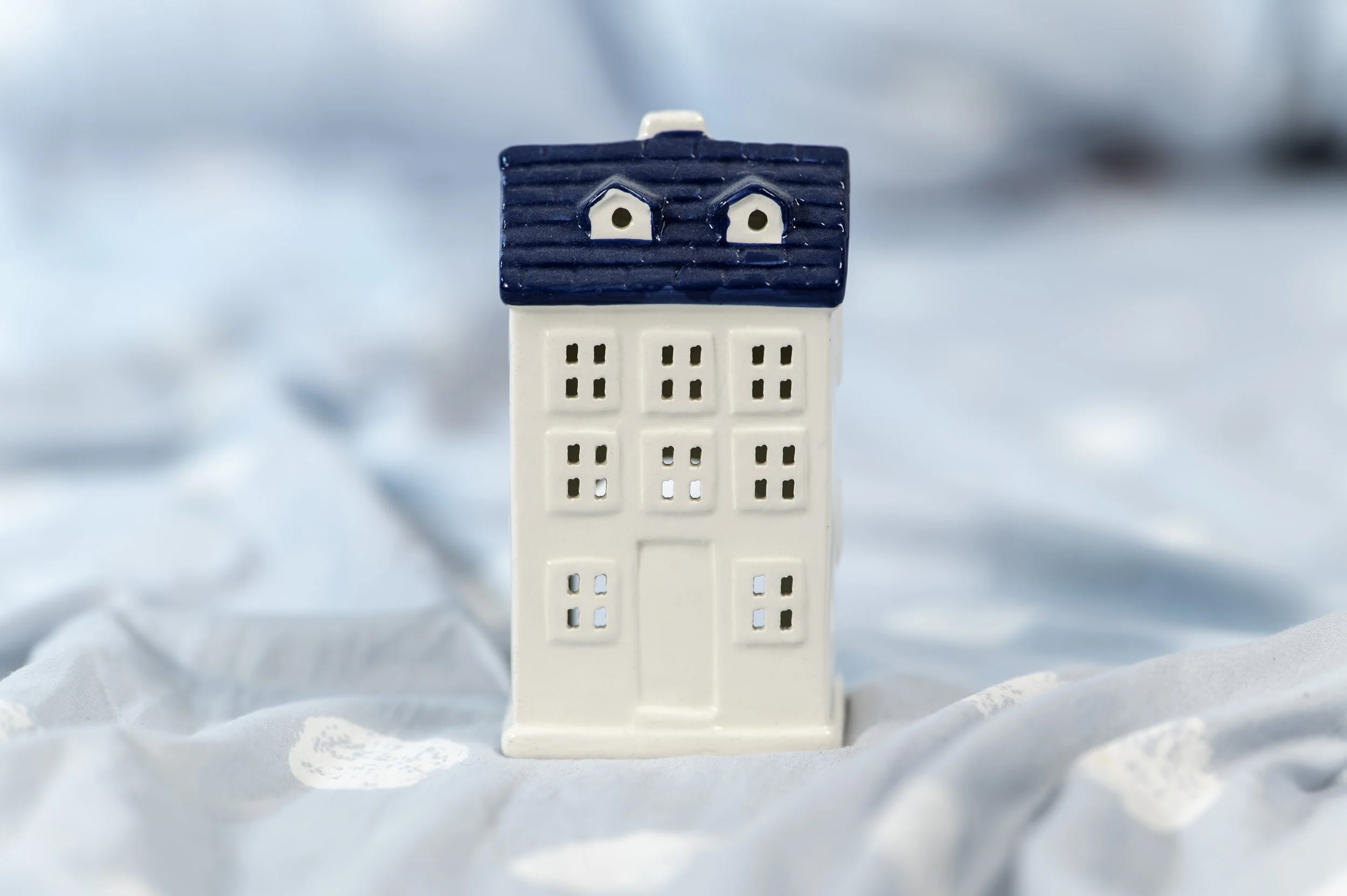 A small ceramic house with a blue roof and white walls, set on a soft, light blue fabric background.