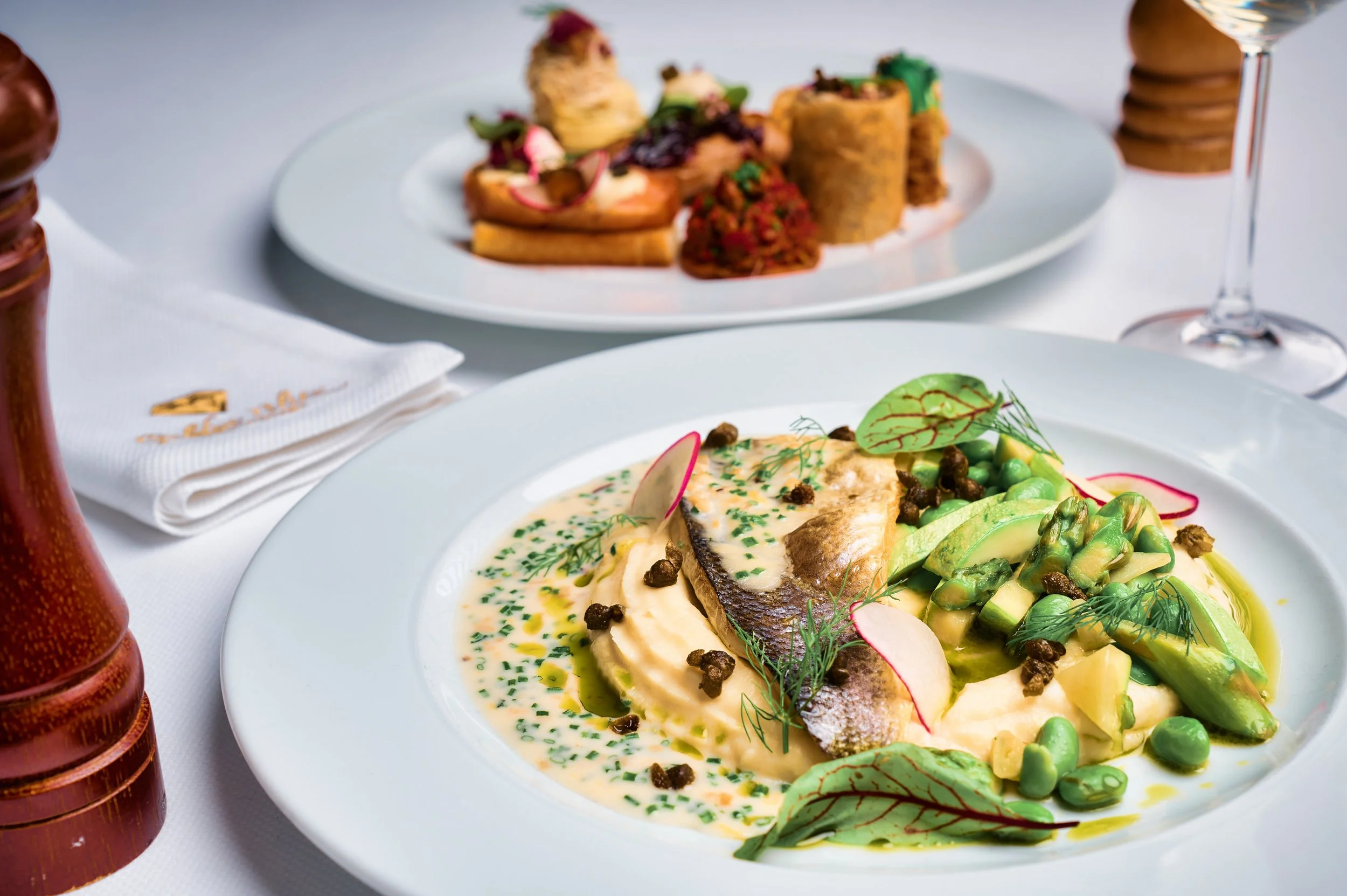 A plated meal with fish fillet topped with herbs and black pepper, accompanied by sliced avocado, green peas, and radish slices, served in a creamy sauce garnished with dill. In the background, a dish with rolled tortillas and a mixture of vegetables