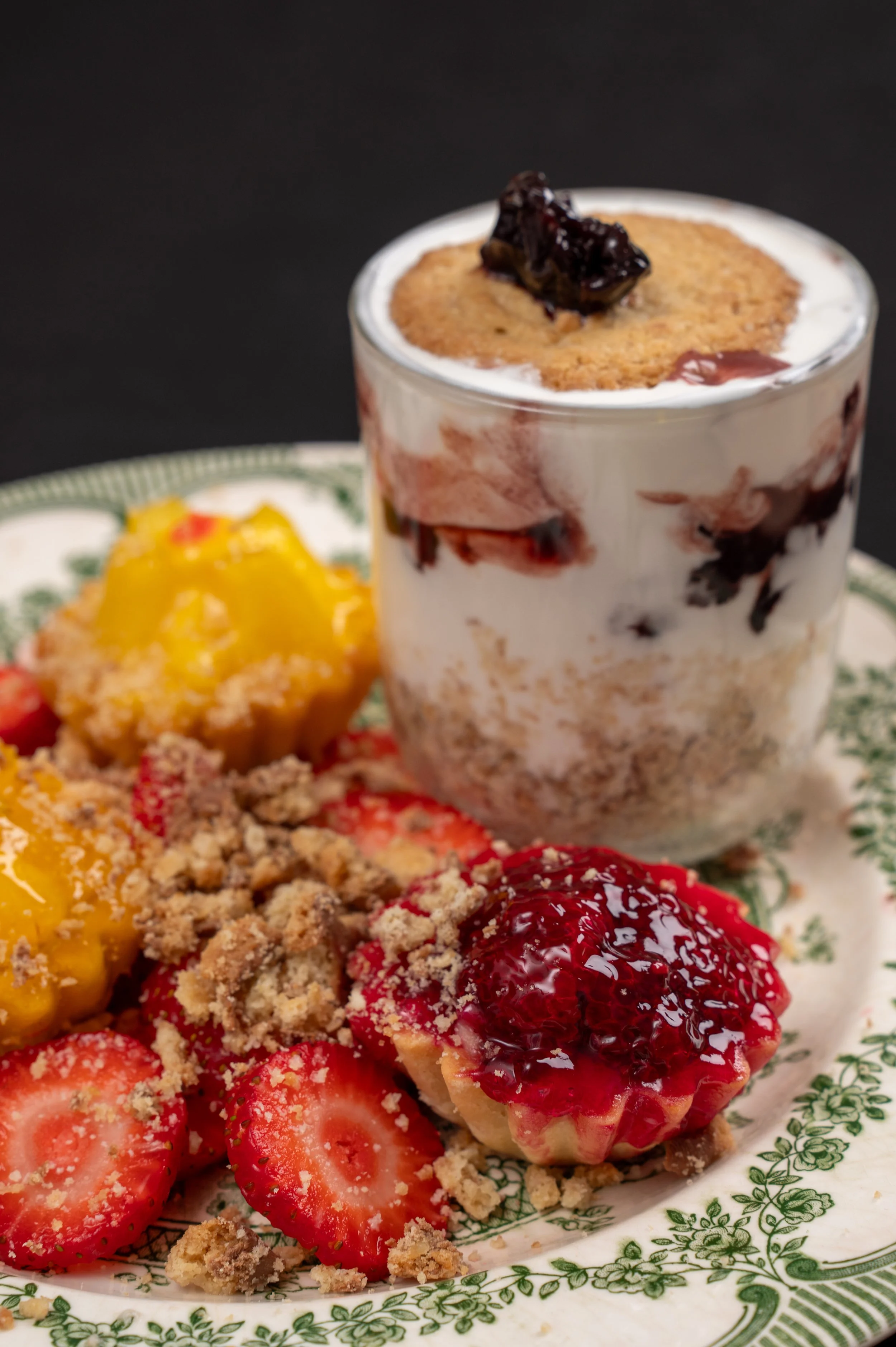 Close-up of a dessert parfait in a glass with layers of whipped cream, cookie crumble, and berry topping, garnished with a cookie on top. On a decorative plate, there are fresh strawberries and slices of strawberries with crumb topping and berry jam.