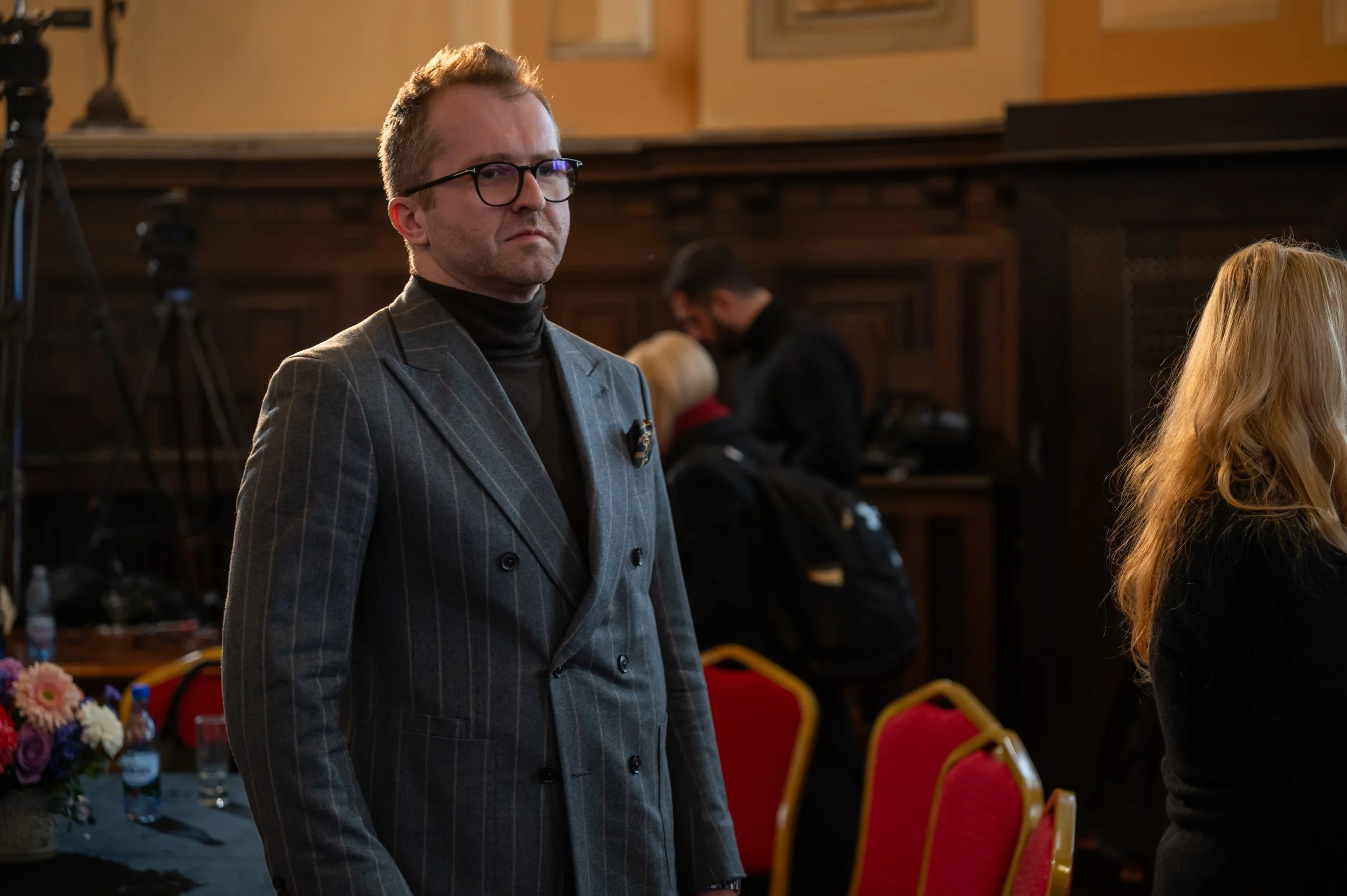 A man with glasses wearing a pinstripe suit standing in a room with wooden walls and red chairs, with several people in the background.