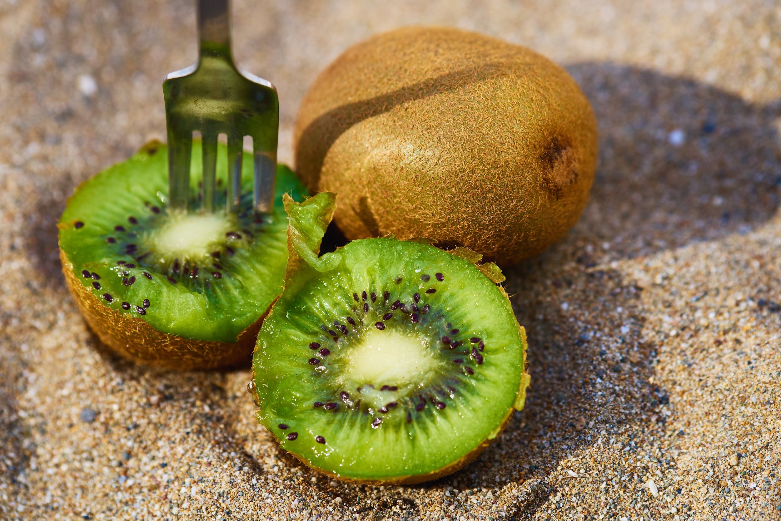 A whole kiwi fruit and a sliced kiwi on sandy ground, with a fork inserted into the sliced kiwi.