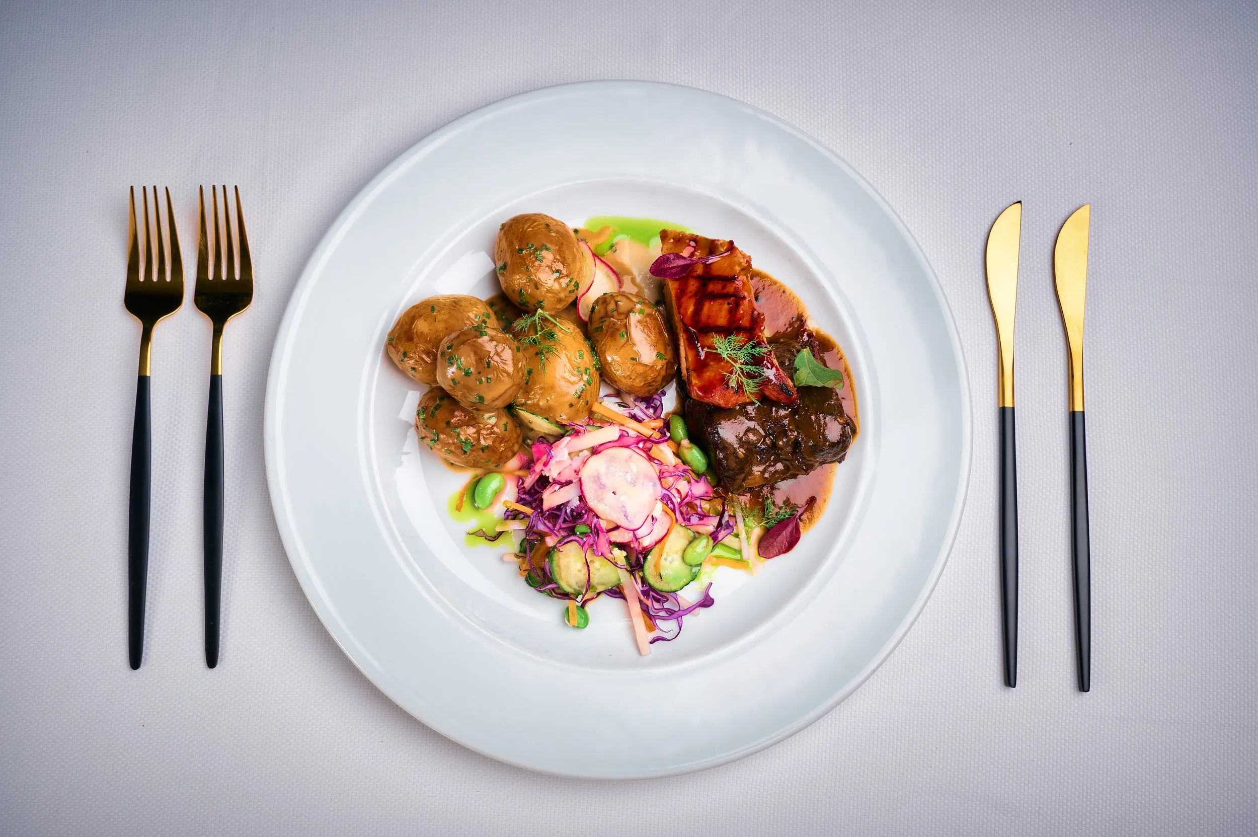 A white plate with roasted potatoes, glazed meat with sauce, and a side salad with vegetables and herbs, served on a white tablecloth with gold and black cutlery.