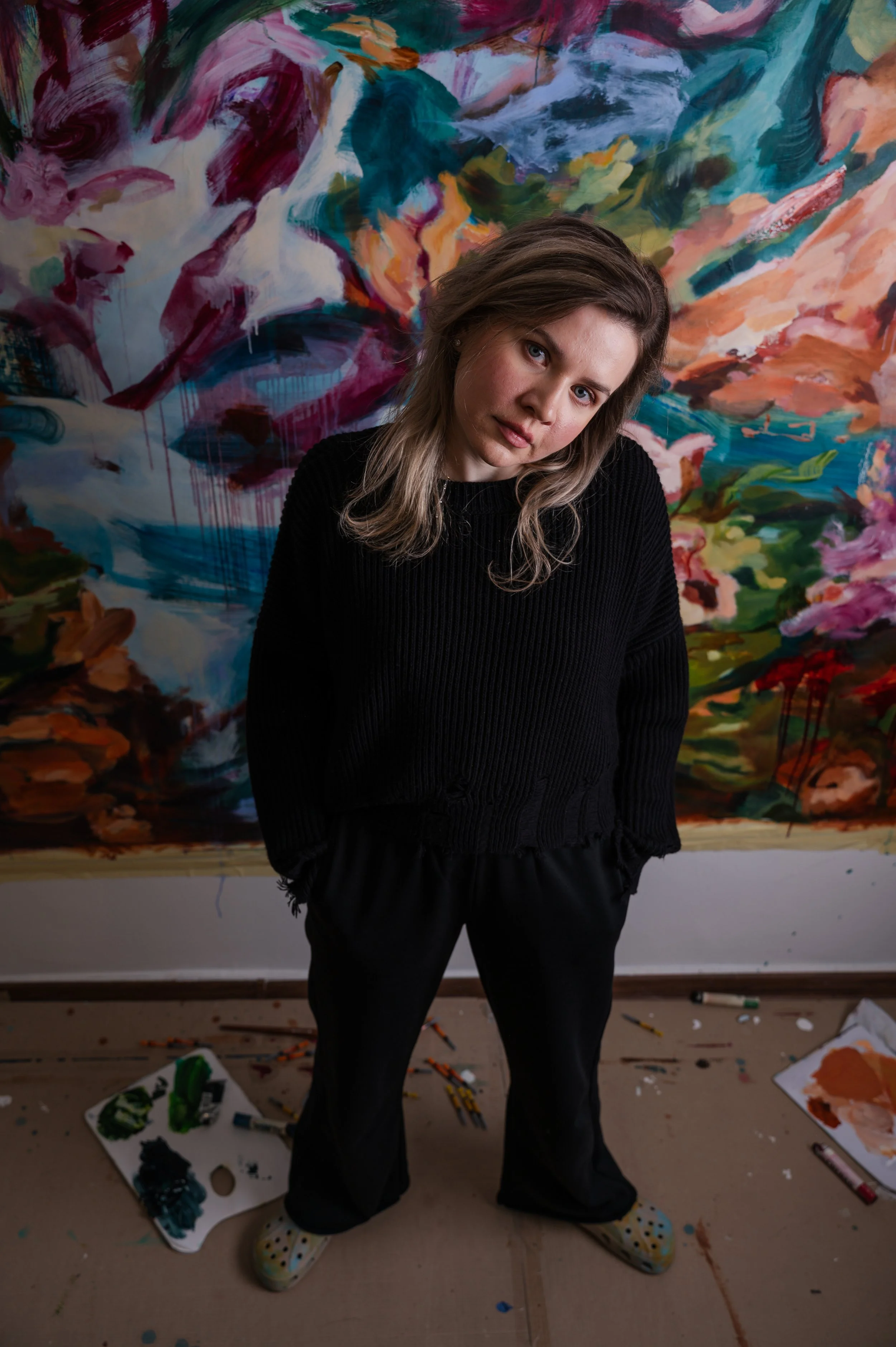 A woman standing in front of a colorful abstract painting. She has shoulder-length brown hair, blue eyes, and is wearing a black ribbed sweater, black pants, and polka dot shoes. The floor around her has art supplies, paint splatters, and a paint pal