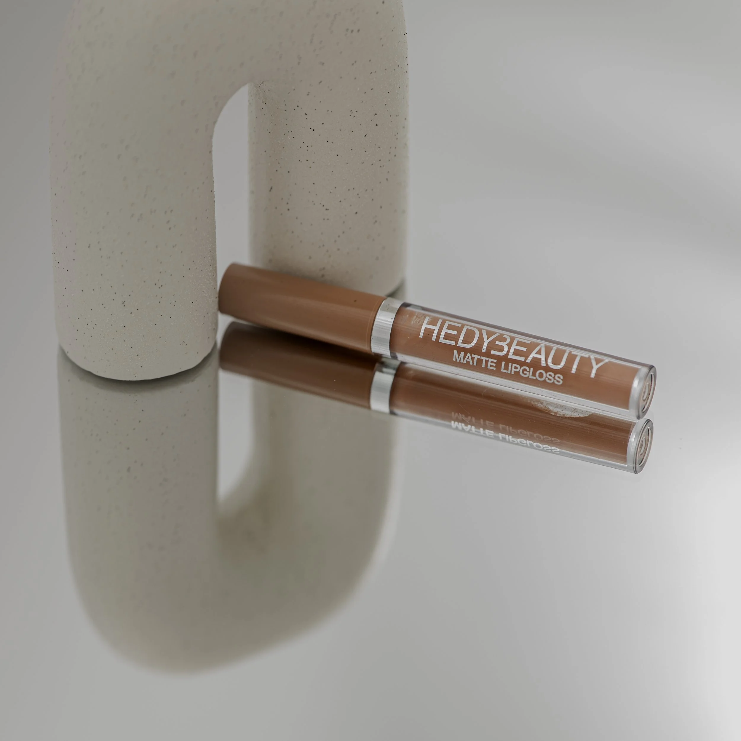 Close-up of a lip gloss tube labeled 'HEDY BEAUTY MATTE LIPGLOSS' lying on a white surface near a speckled beige object.