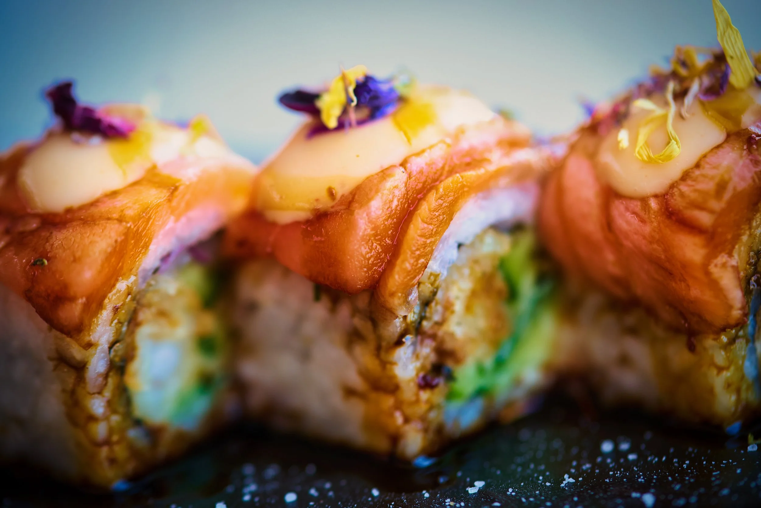 Close-up of sushi rolls topped with sauce, edible flowers, and colorful ingredients.