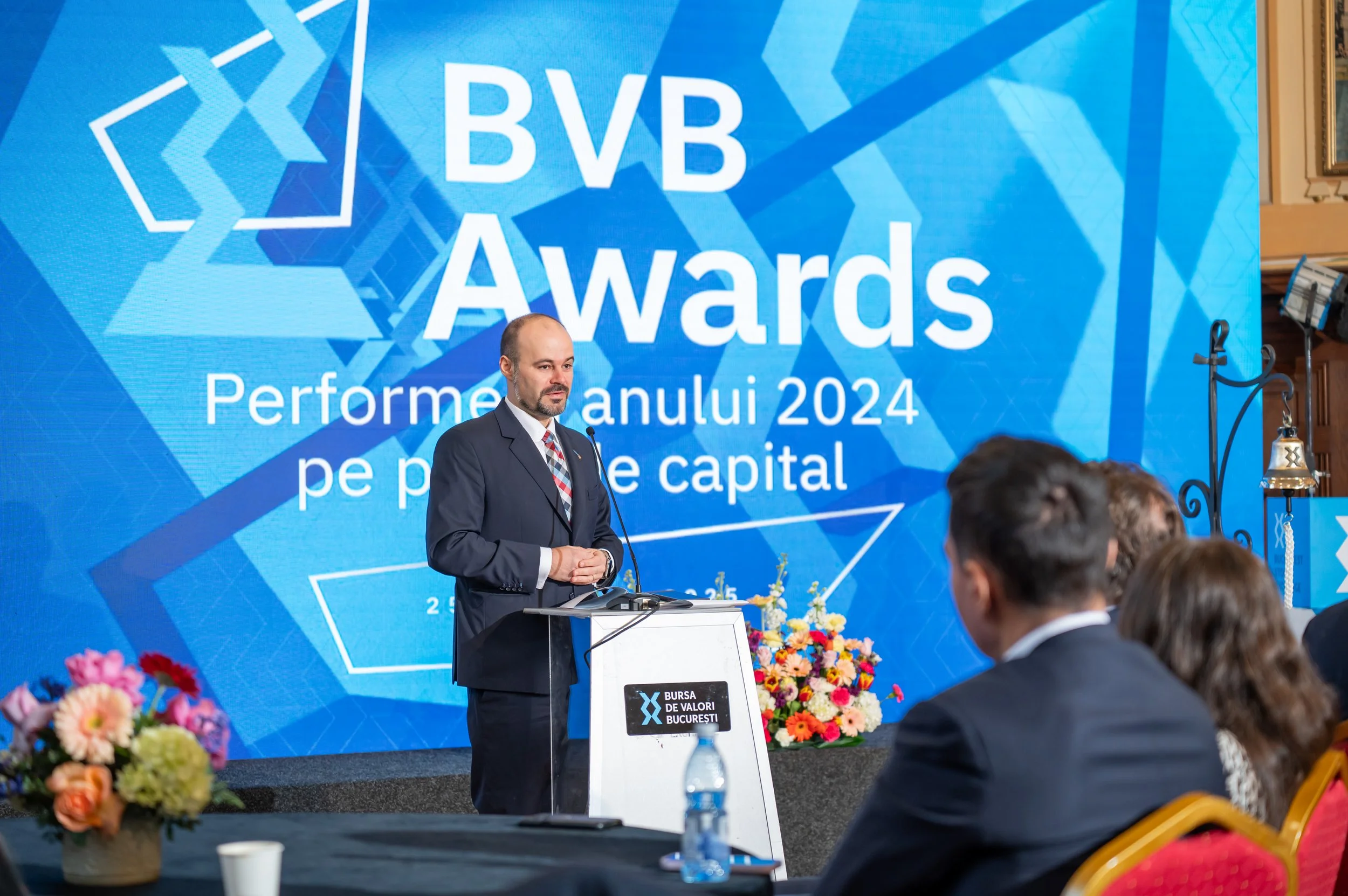 A man in a suit speaking at a podium during the BVB Awards event in Bucharest 2024, with a large blue backdrop displaying the event name and date. Audience members and floral arrangements are visible in the foreground.