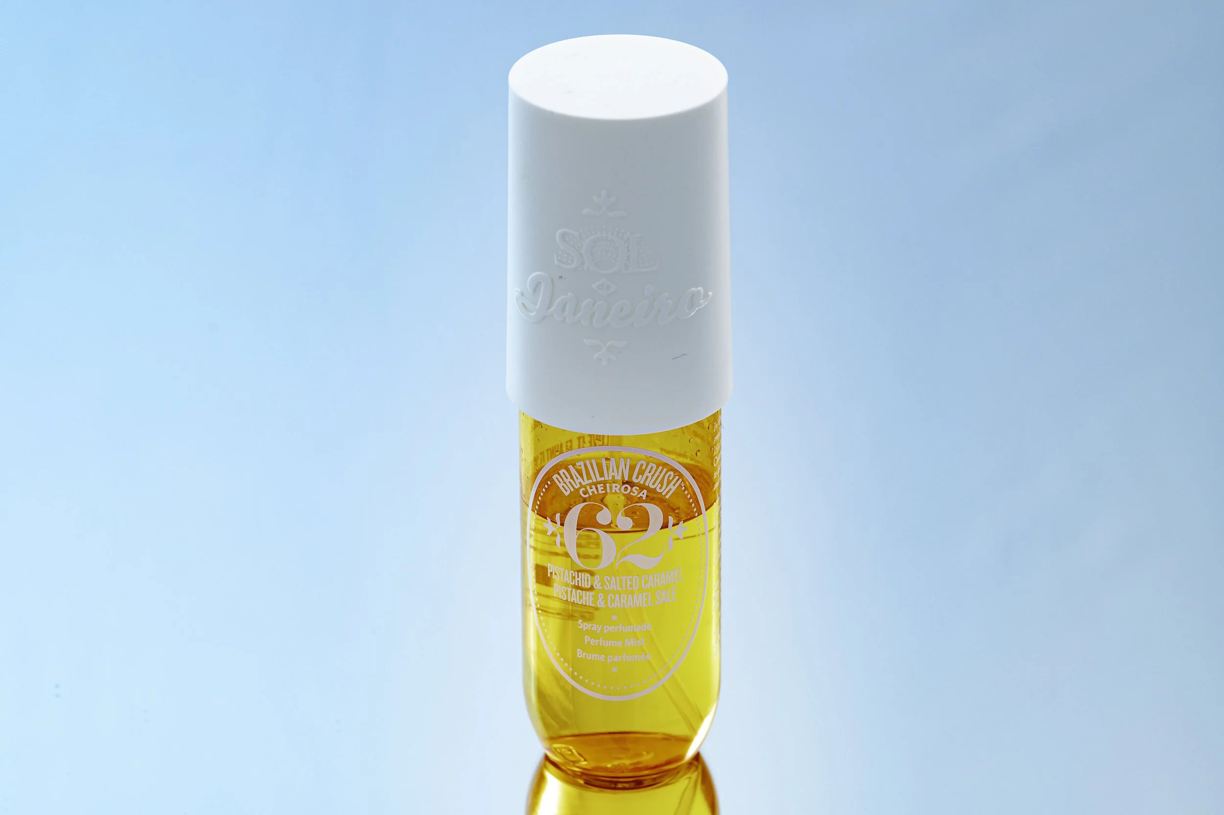 A spray perfume bottle with an orange and yellow label that reads 'Brazilian Crush Cheirosa 62 Pistachio & Salted Caramel' against a light blue background.