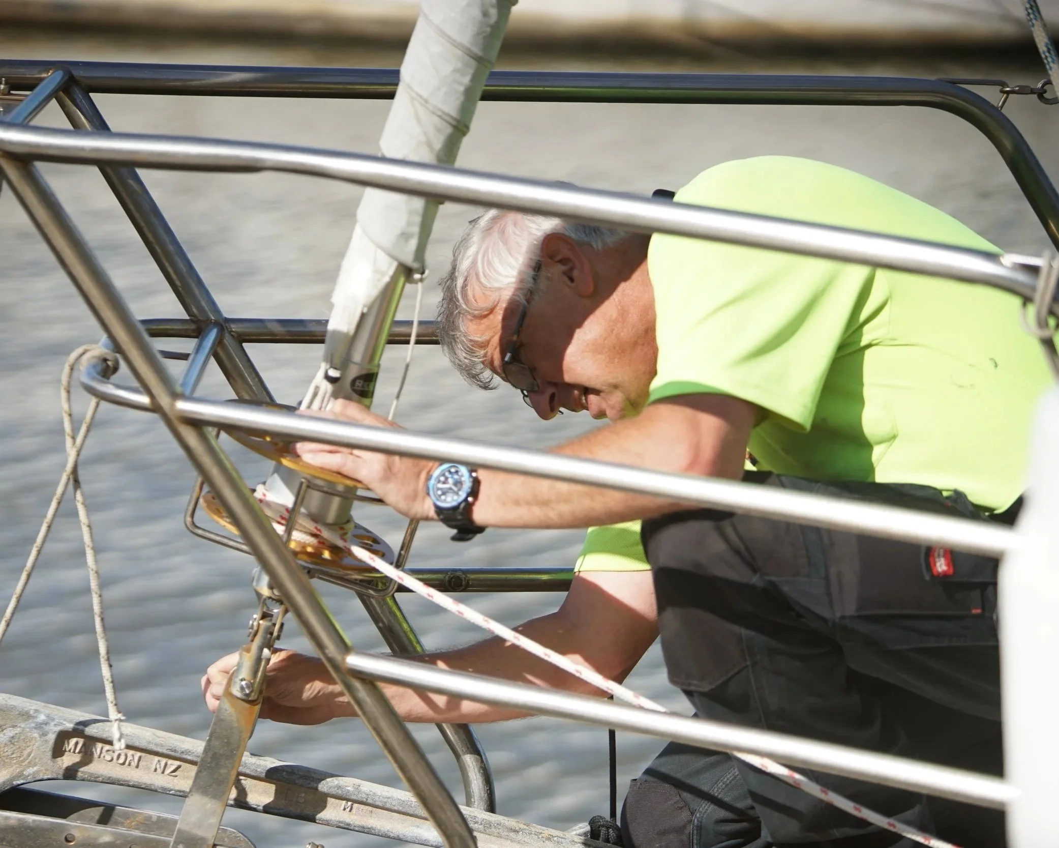 Close-up of a man adjusting equipment on a small sailboat, wearing a neon green shirt, a watch, and glasses, with water in the background.
