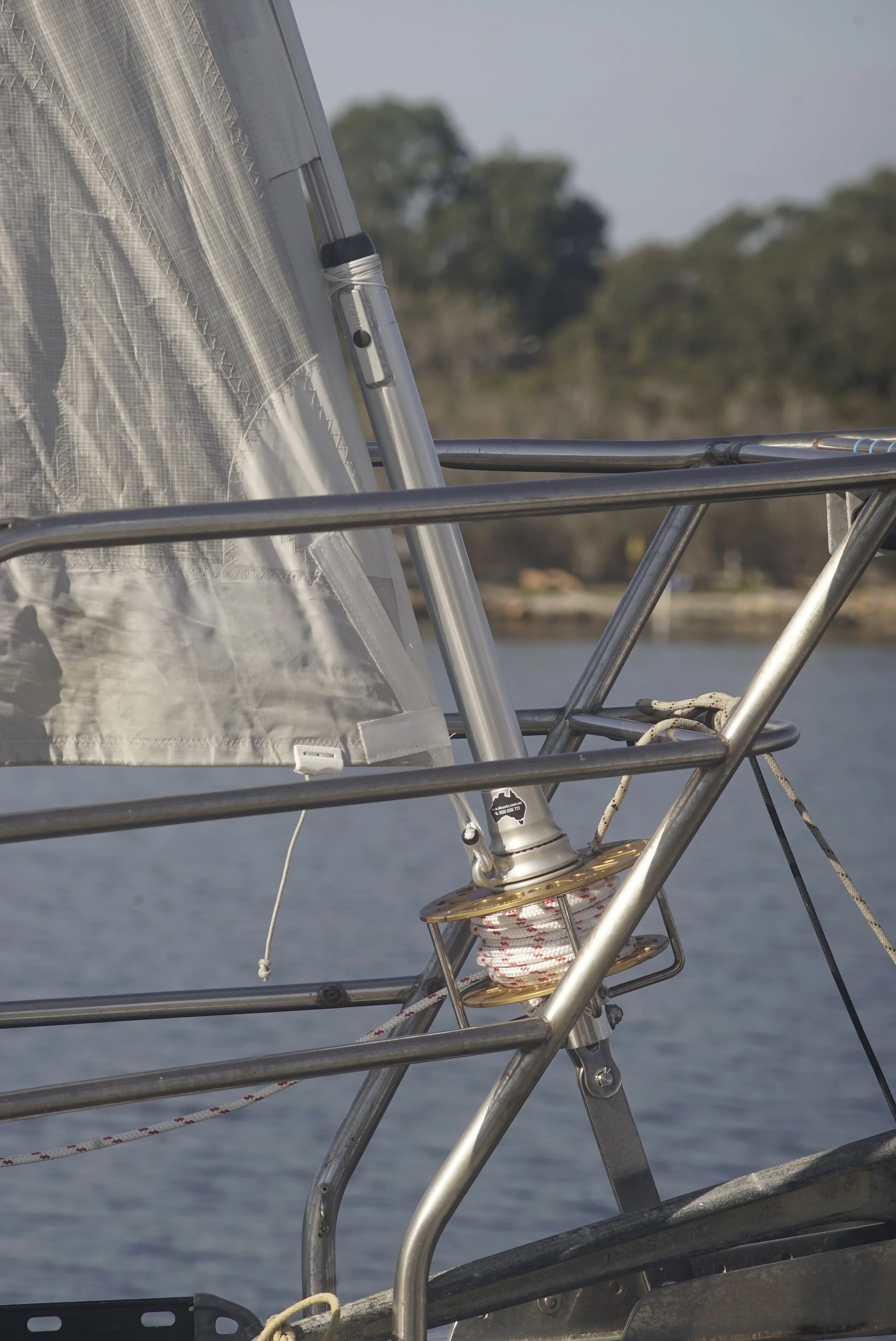 Close-up view of a sailboat's rigging and sail with ropes and metal framework, on water with blurred trees in the background.