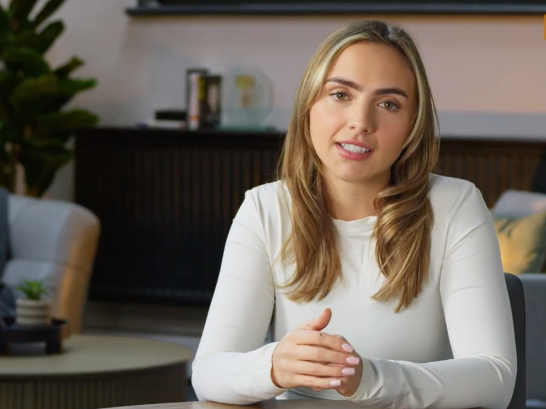 A woman with long blonde hair and light skin, wearing a white shirt, sitting at a table in a modern room, answering questions in a recorded talking-head interview.