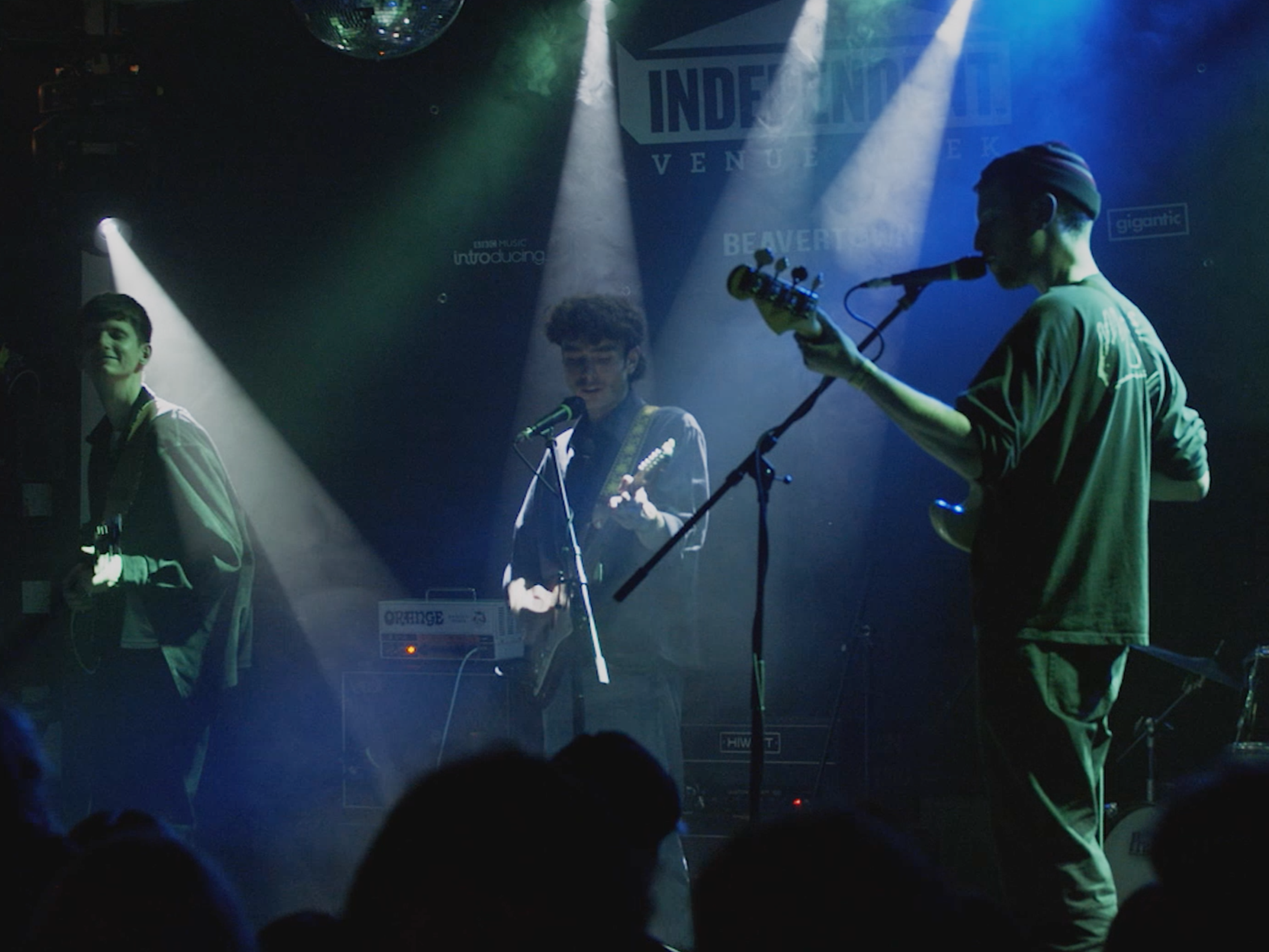 Band performing on stage at live music venue, featuring the band playing guitars and singing into microphones under spotlights seen through the lens of the camera.