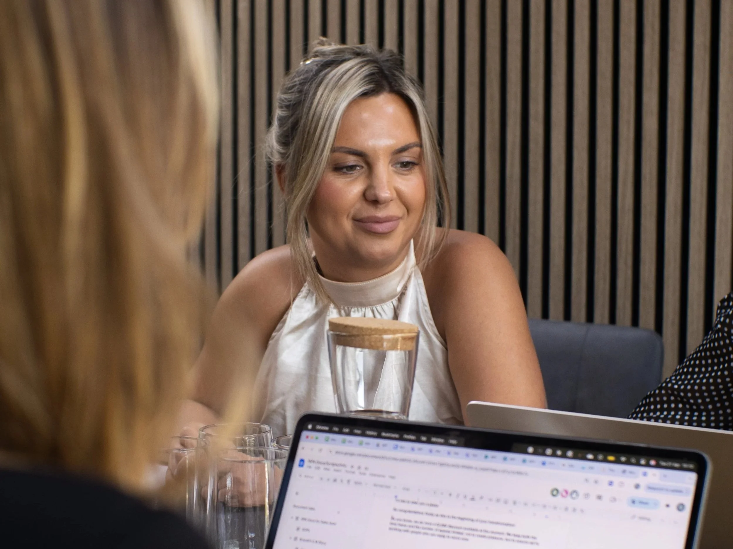A woman with blonde hair and a light-colored top is sitting at a table, smiling softly. There are glasses and a laptop with an open webpage in front of her. The background features a wooden slat wall.