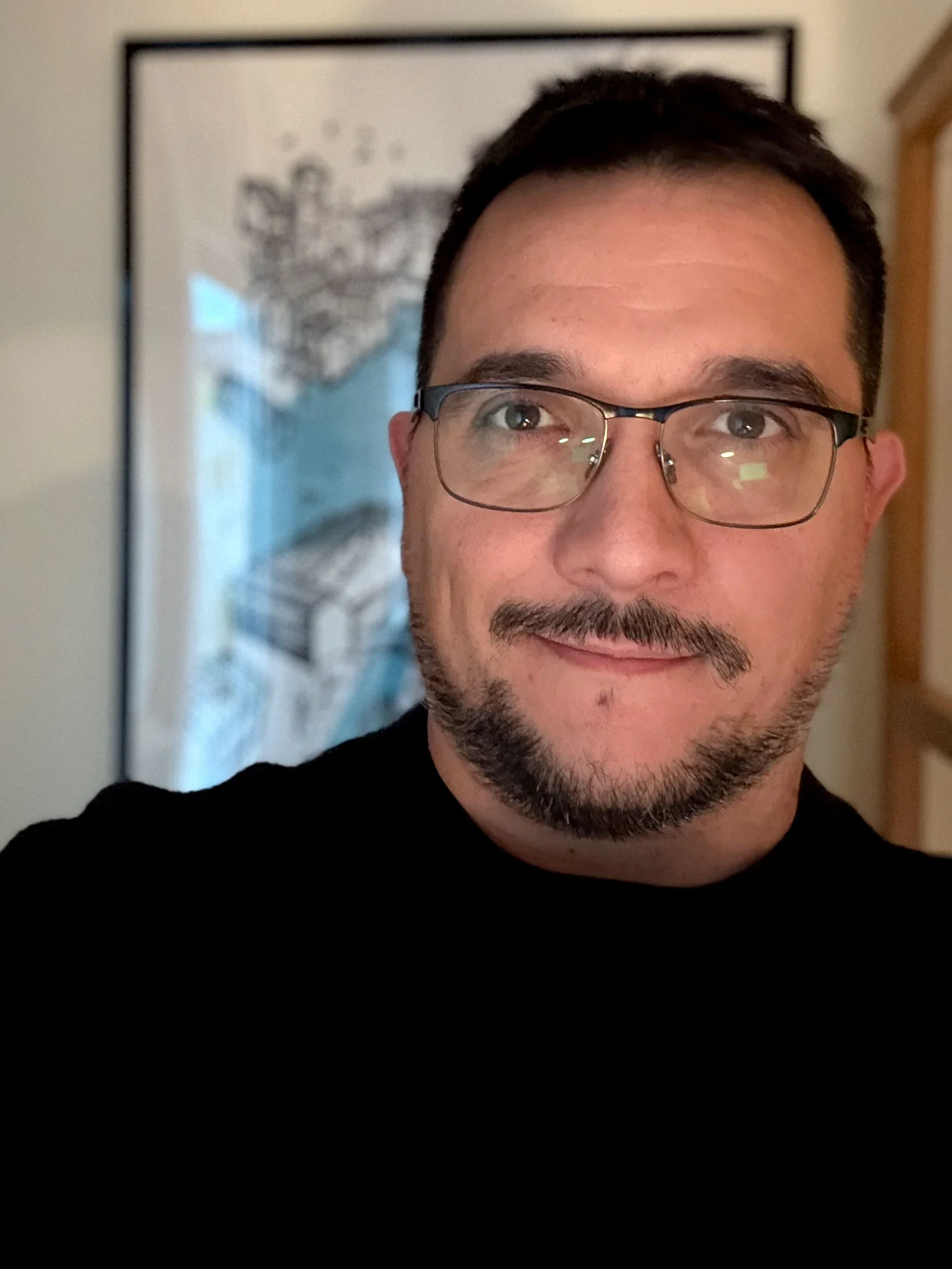 Ritratto fotografico di Davide Romagnoli, illustratore e graphic designer freelance, con occhiali, capelli corti e barba. Alle sue spalle è appesa una delle sue illustrazioni, stampata e incorniciata.