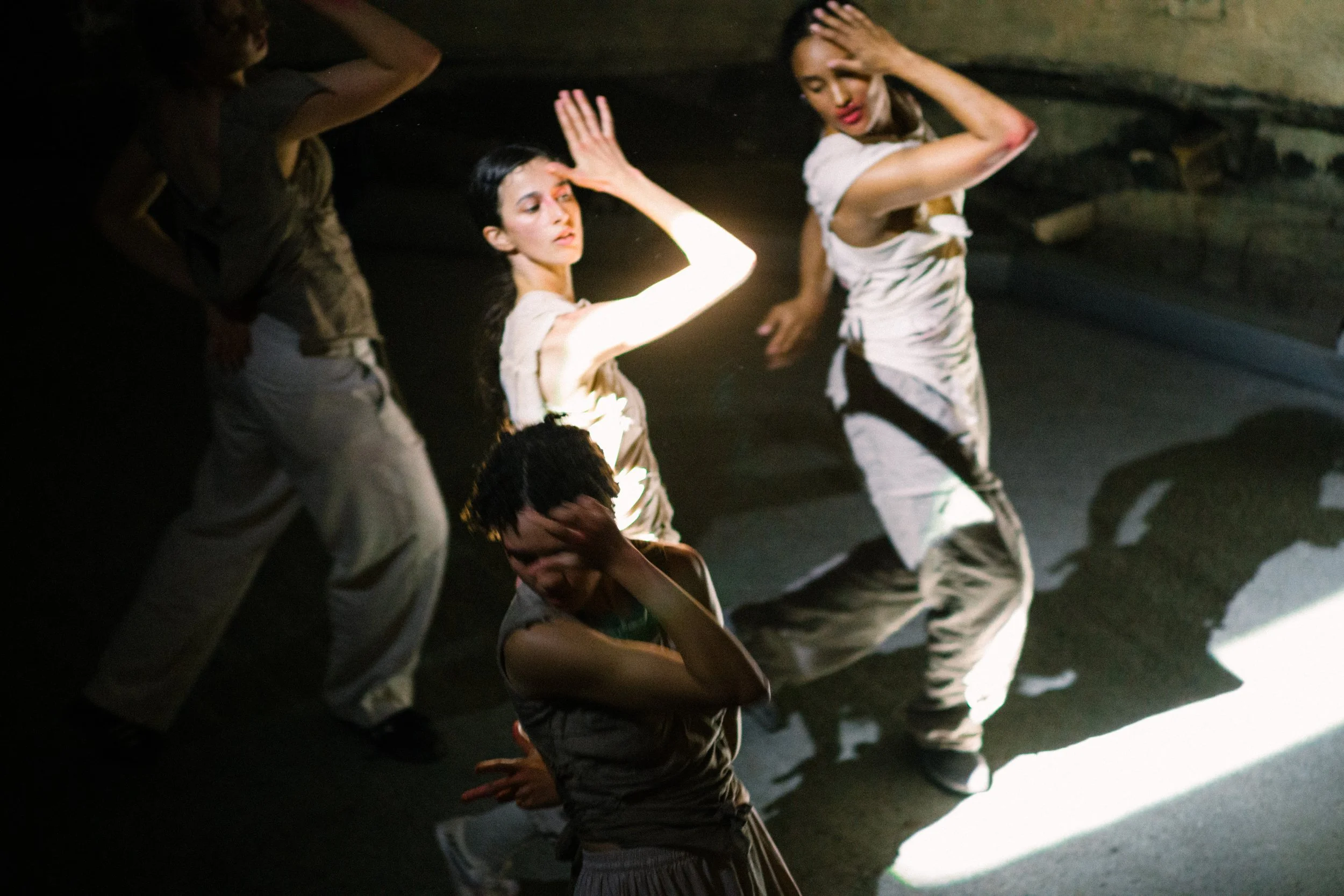 A group of young women and girls dancing in a dimly lit room, some with hands on their heads, one with eyes closed, under a focused light creating shadows on the floor.