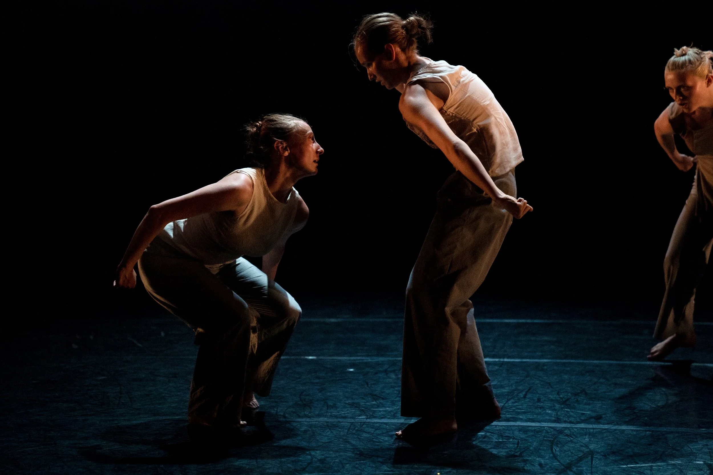 Three female dancers performing on stage in dim lighting, wearing casual clothing, with one bending down and another standing while engaging in a dance movement.