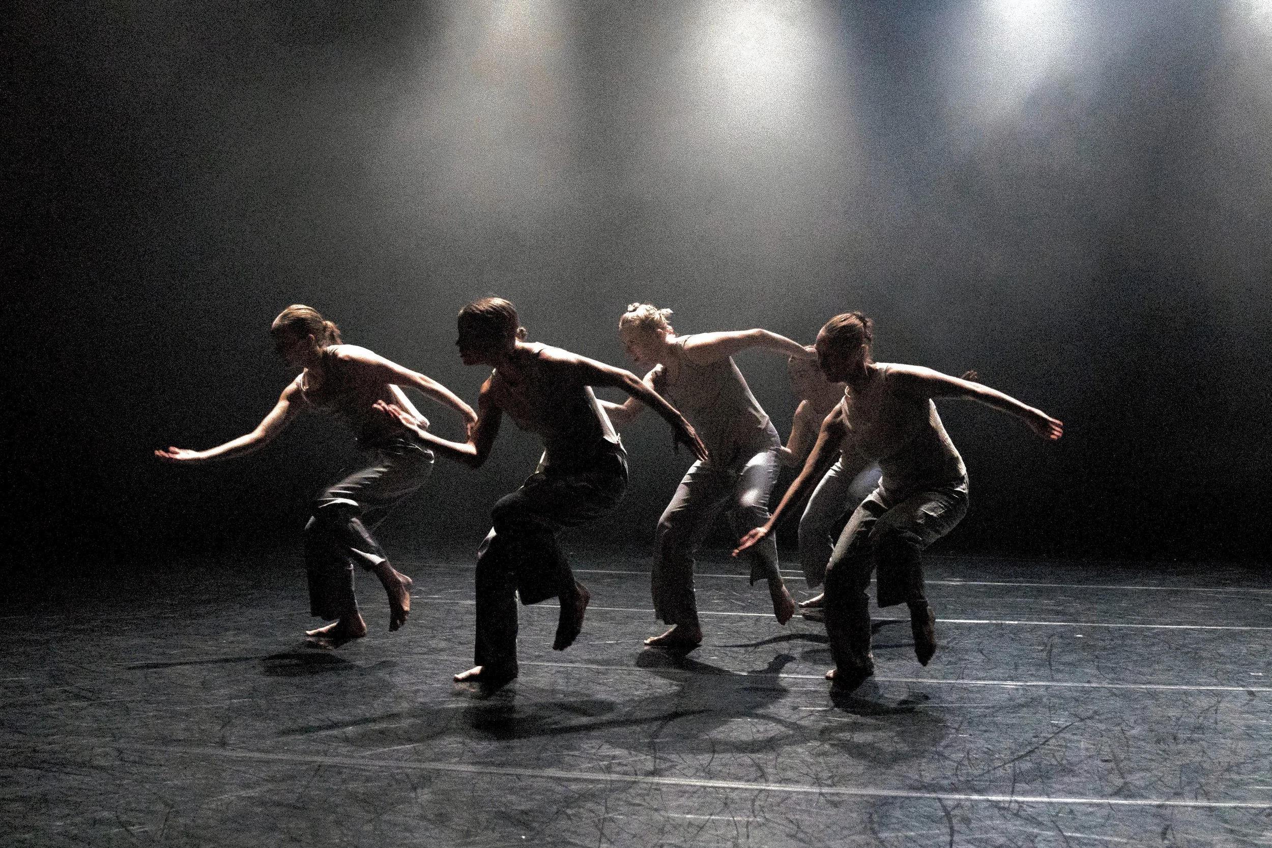 A group of five dancers in white clothing performing on a dark stage with dramatic lighting.
