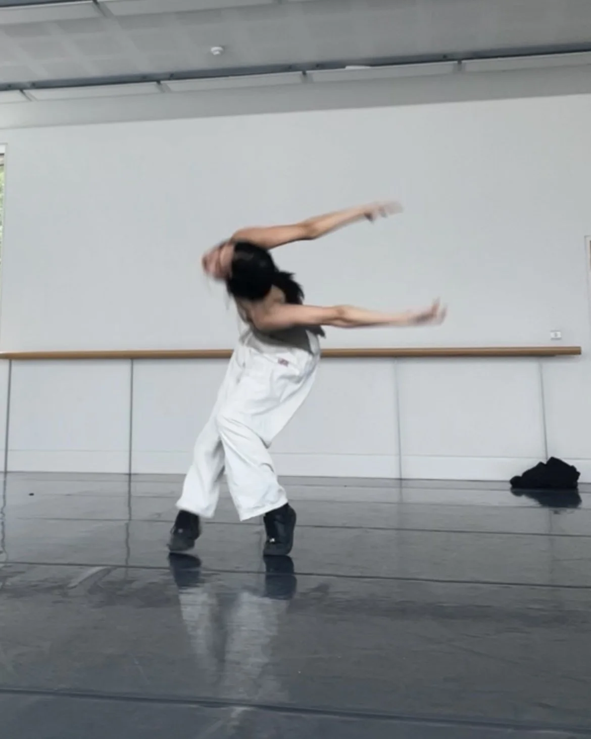 A person dressed in white pants and black shoes performing a dance move in a dance studio with a black floor, white walls, and a wooden ballet barre.