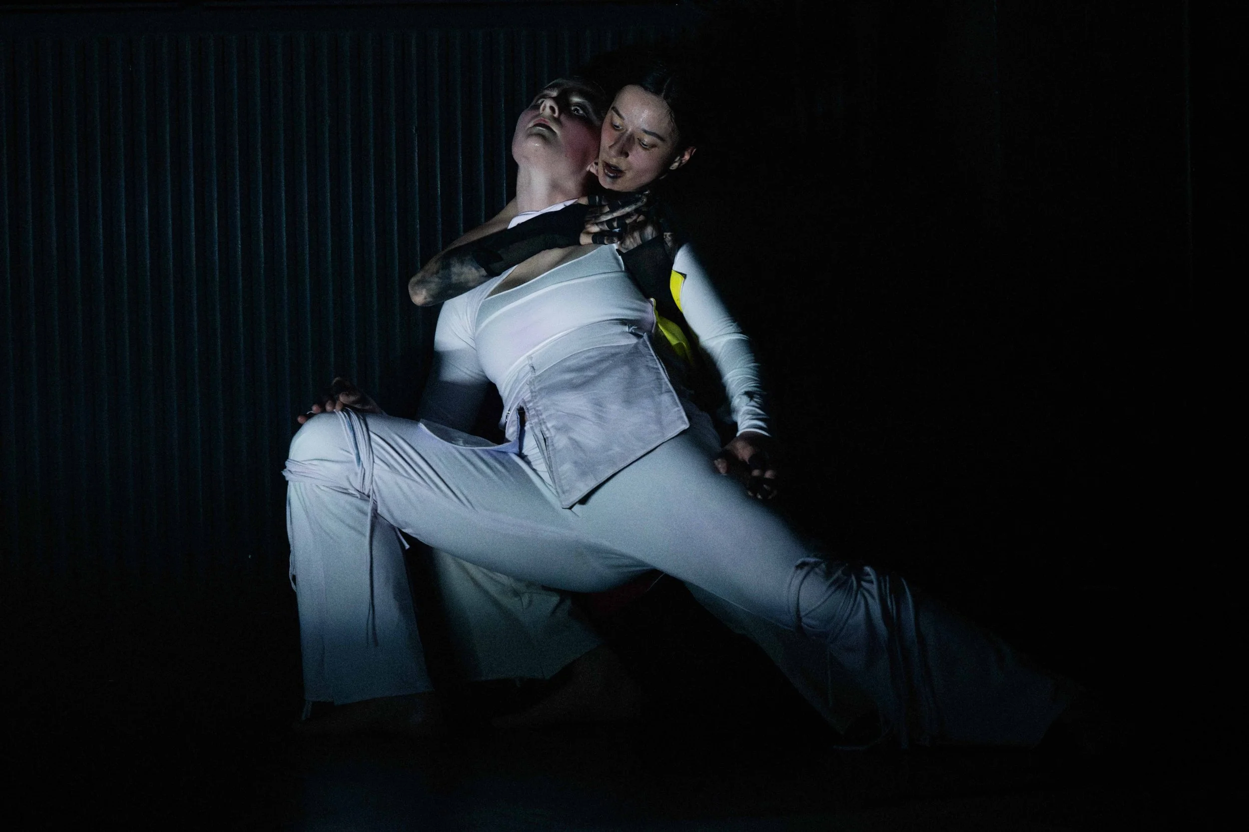 A performer dressed in black and white with bold makeup is embracing another person, who is wearing a black top and white pants, against a dark, textured background.