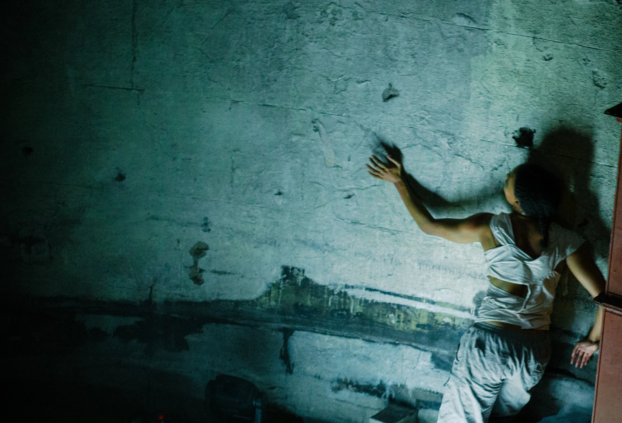 A person doing a handstand against a rough white wall with visible cracks and spots, in a dimly lit room.