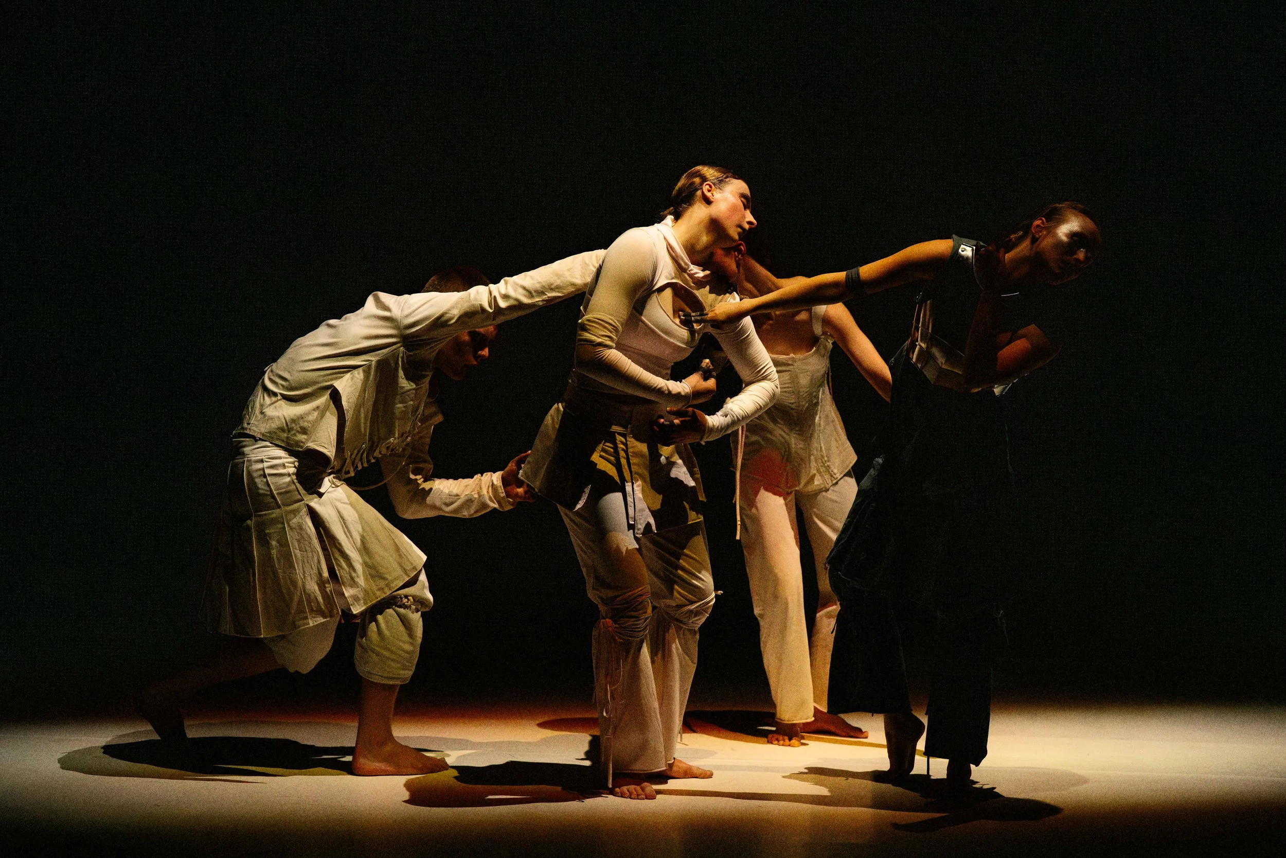 A group of performers in beige and black costumes are linked together at the waist and hands, performing a dance on a dimly lit stage.