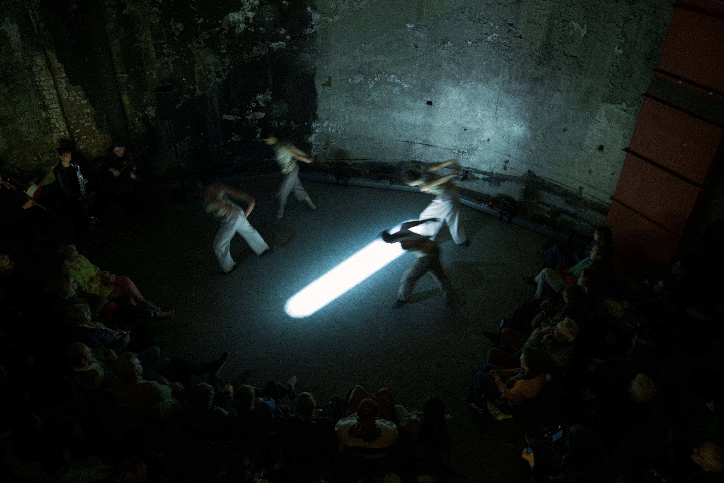 A dark performance space with an audience seated around a small dance performance in the center, where performers are dressed in white and are moving under a beam of light.